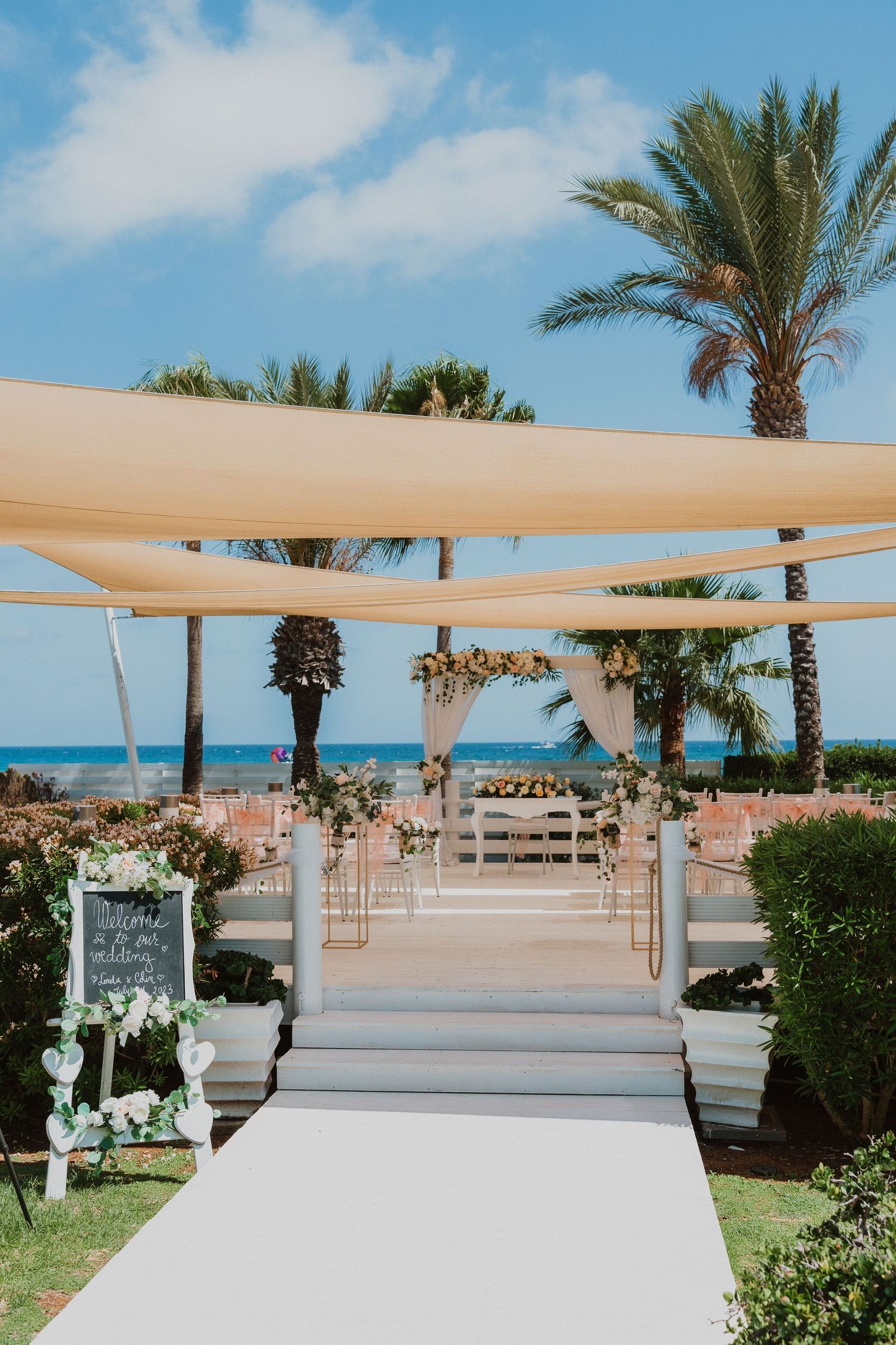 Elegant beachfront wedding ceremony setup with white aisle runner, canopy drapes and palm trees against blue ocean backdrop.