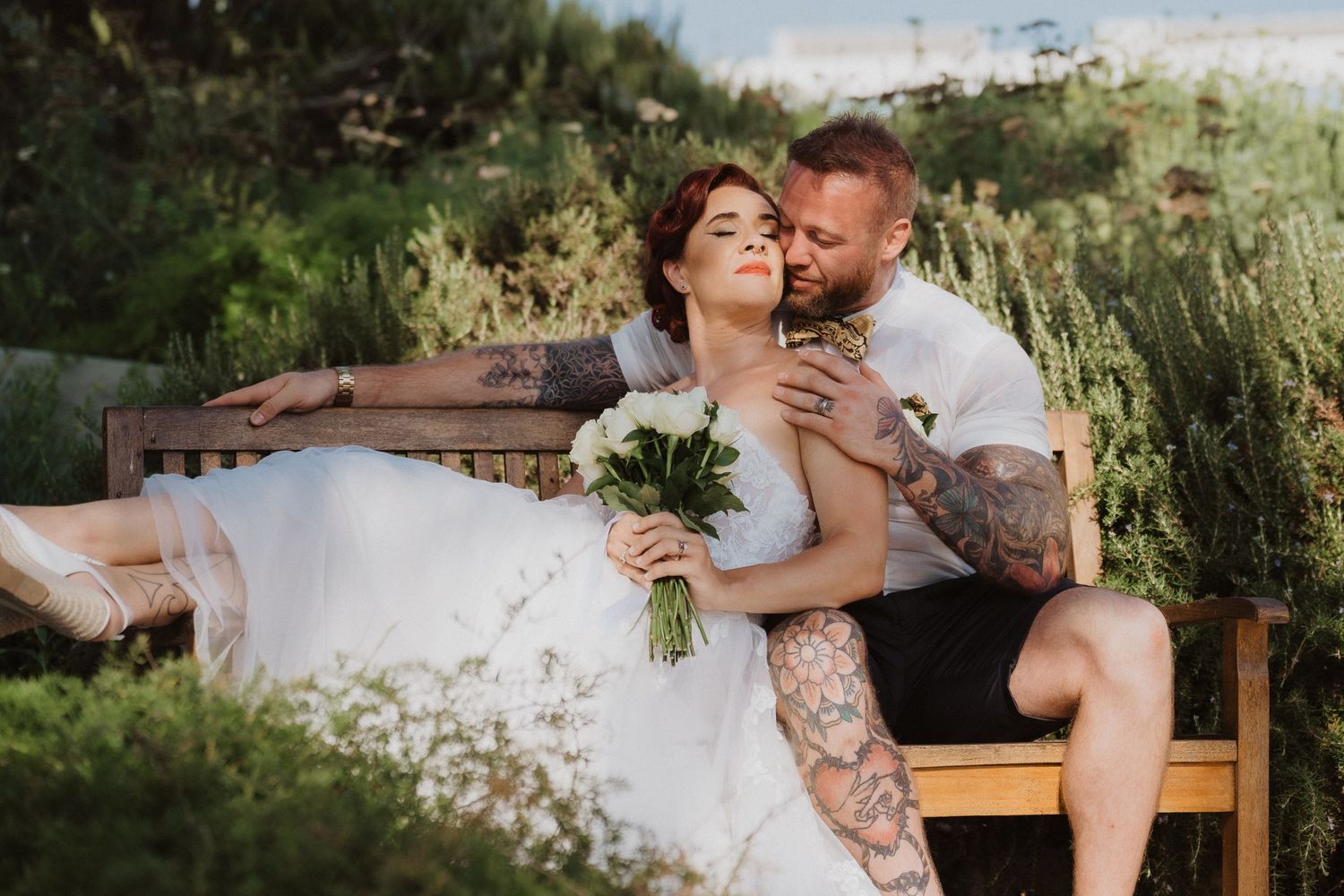 Intimate wedding portrait on wooden bench with white bouquet and garden backdrop.