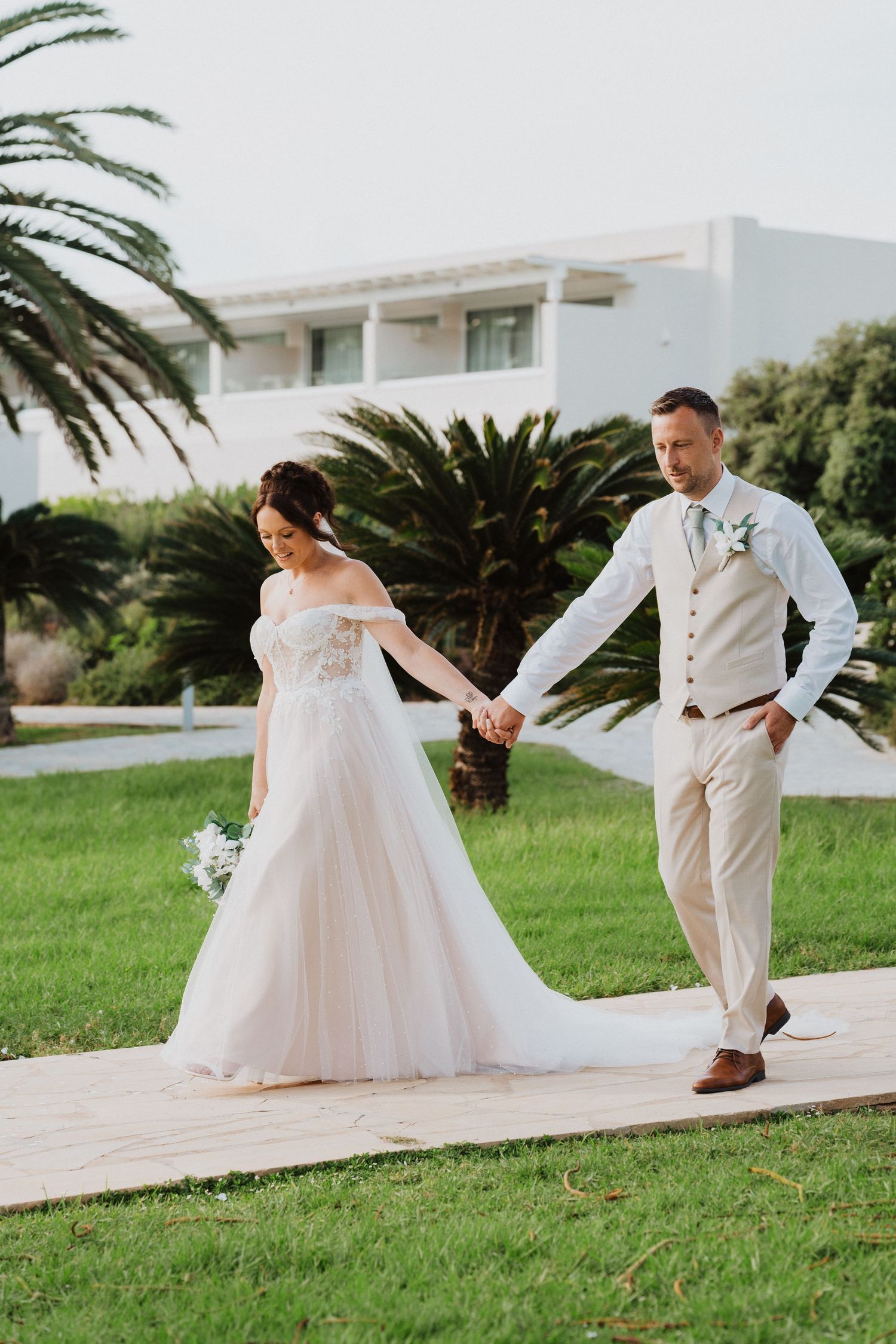 A bride and groom walk hand-in-hand on a paved path with palm trees and a white building in the background.