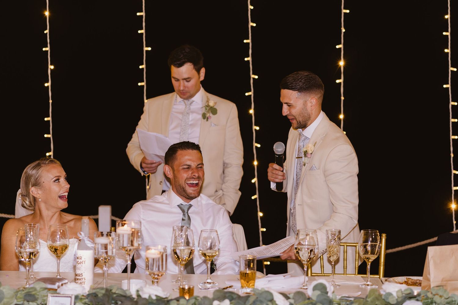 Joyful reactions during wedding reception speeches at decorated head table.