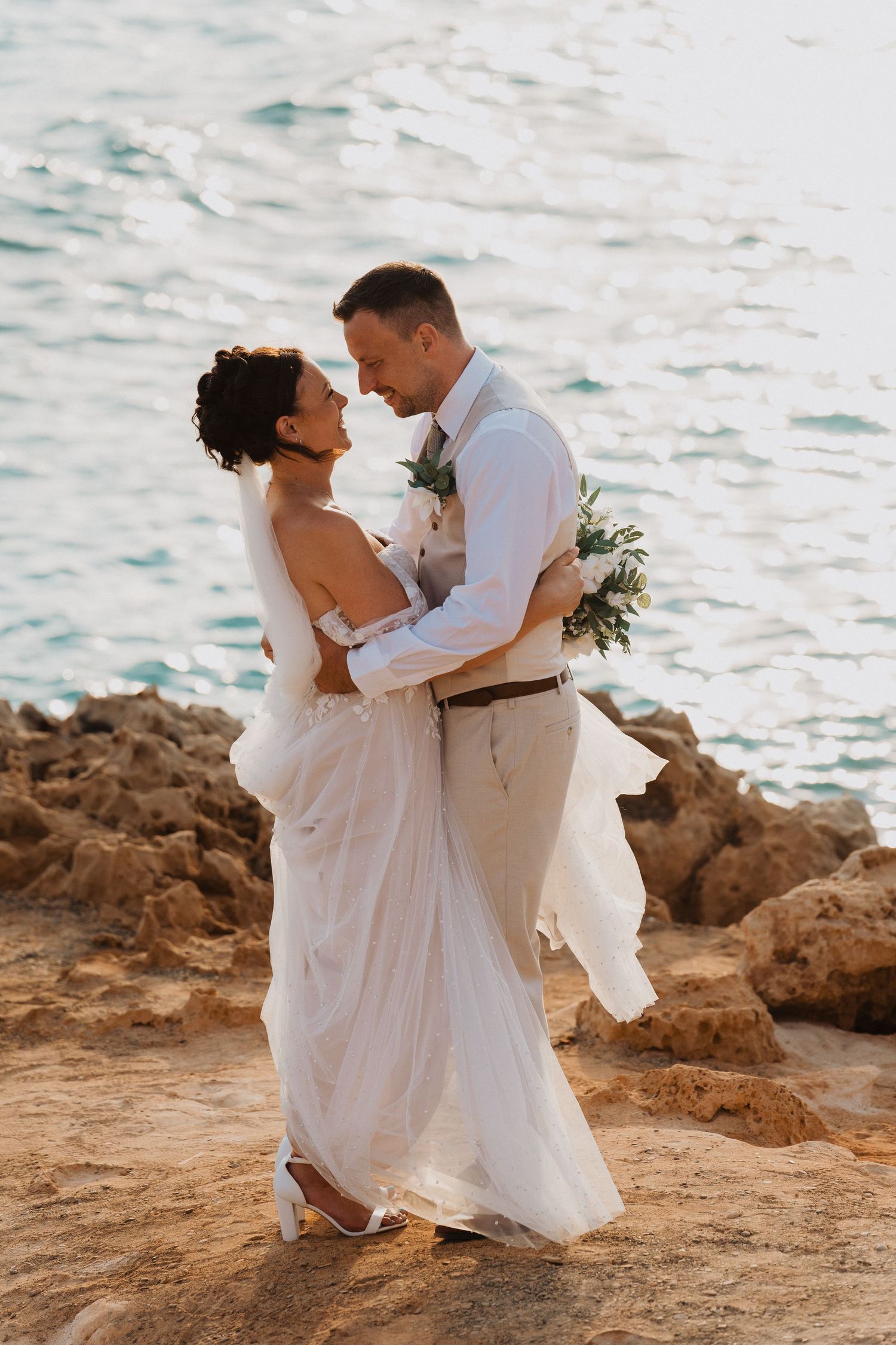 Romantic oceanside wedding couple embraces on rocky cliff during sunset in flowing white dress and casual attire.