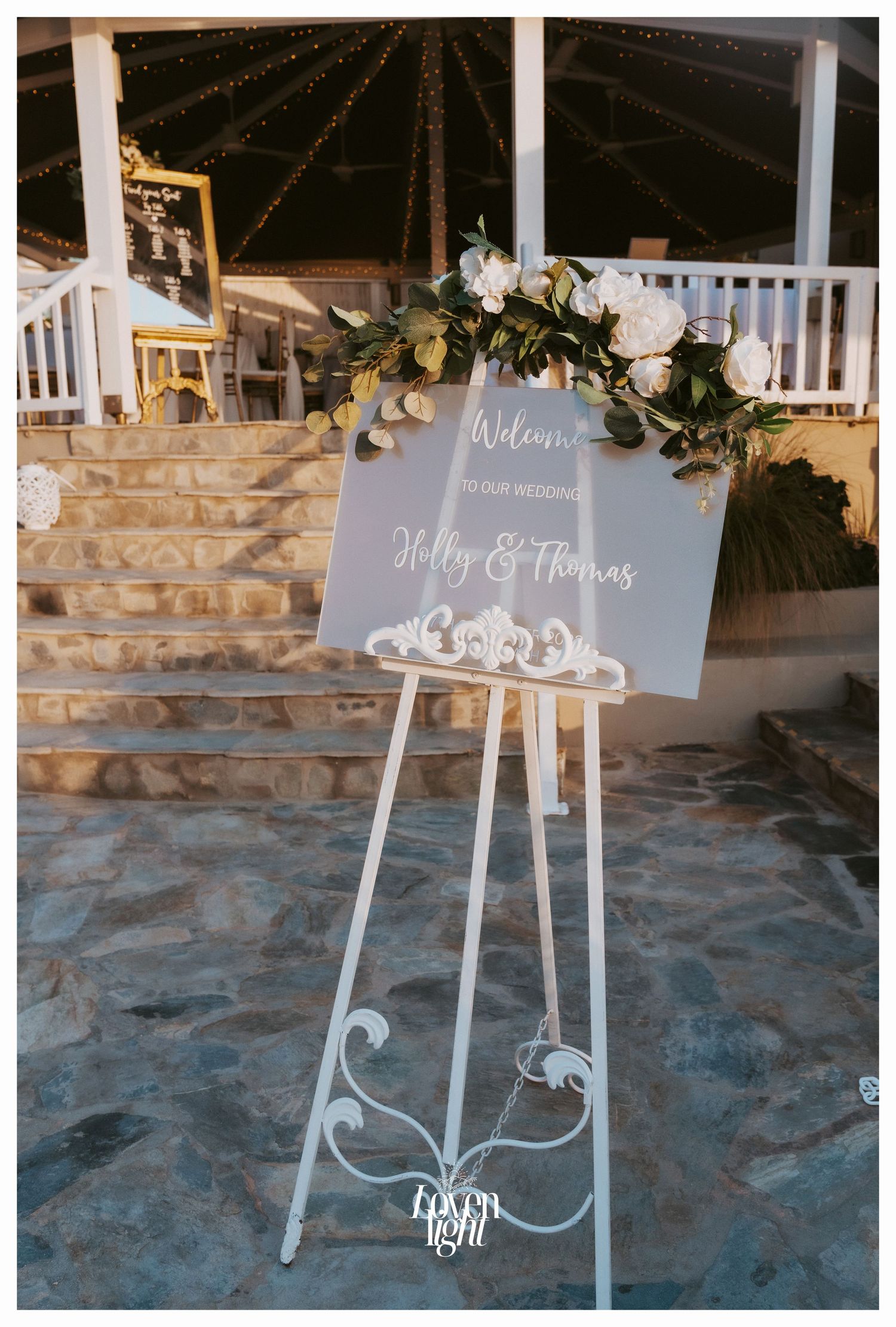 Elegant white wedding welcome sign with floral decorations displayed on an ornate easel at an outdoor venue.
