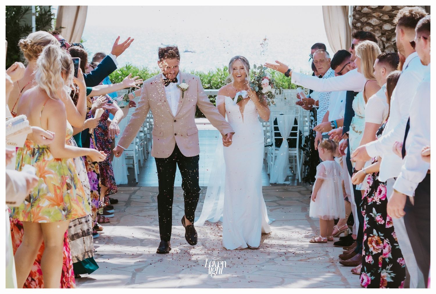 Newlyweds celebrate while walking down an aisle lined with cheering guests in colorful attire.