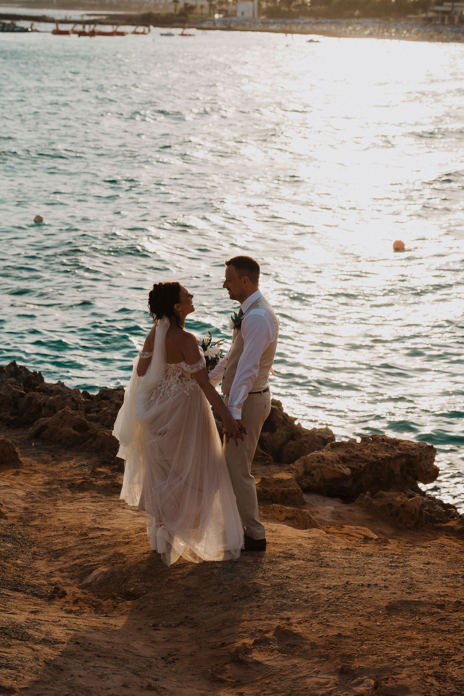 A couple in wedding attire embrace on a rocky cliff overlooking the ocean at sunset.