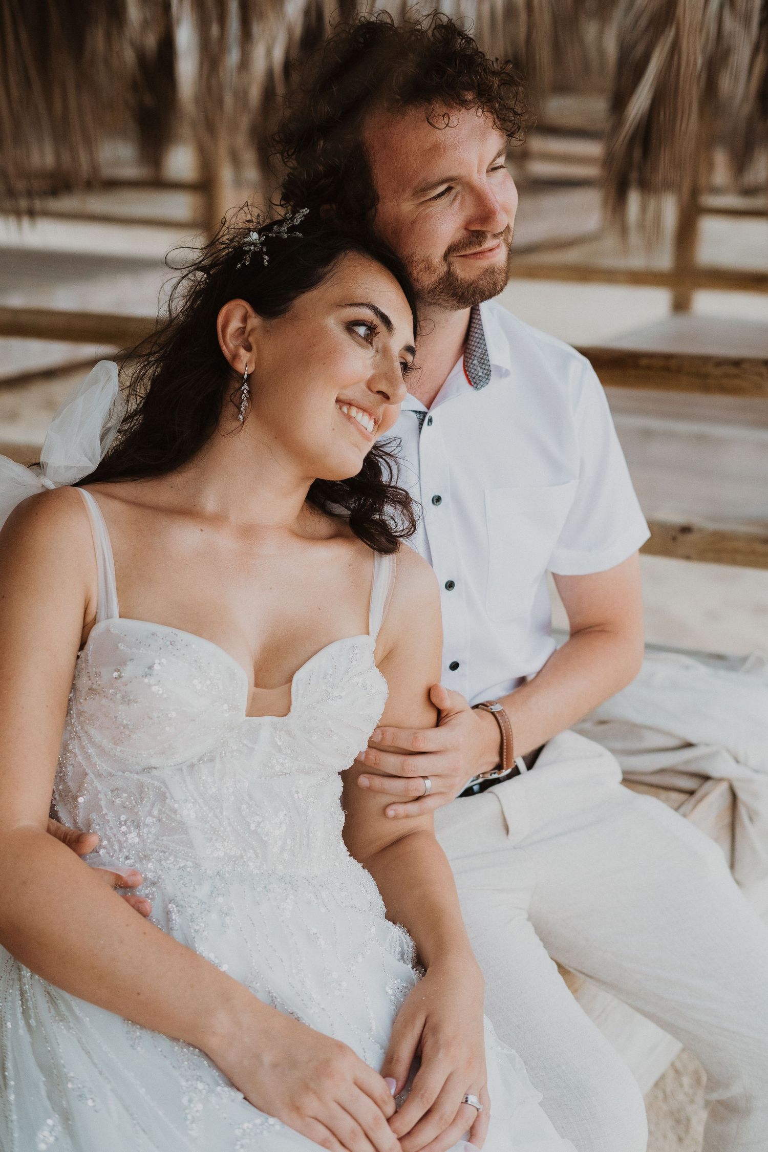 An intimate wedding portrait shows a couple in white sharing a tender moment under palm fronds.