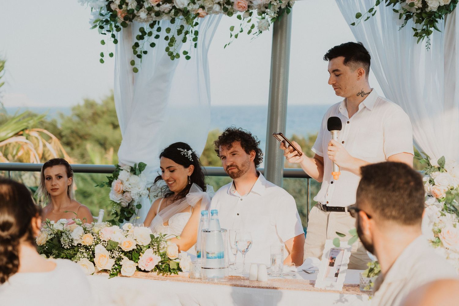 Wedding reception speech moment under floral-decorated white canopy by the sea.