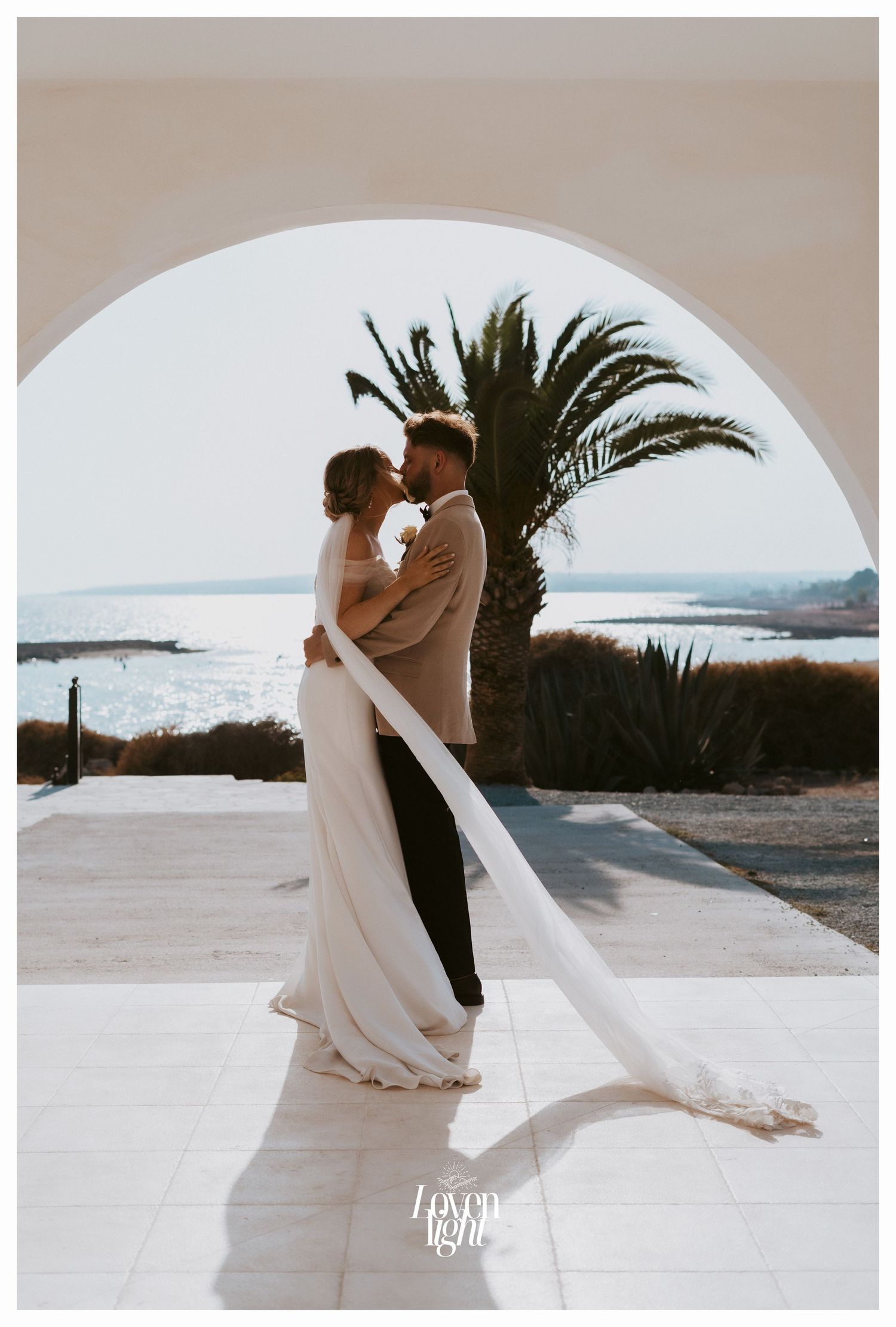 Intimate wedding moment captured through archway with palm tree and Mediterranean seascape.