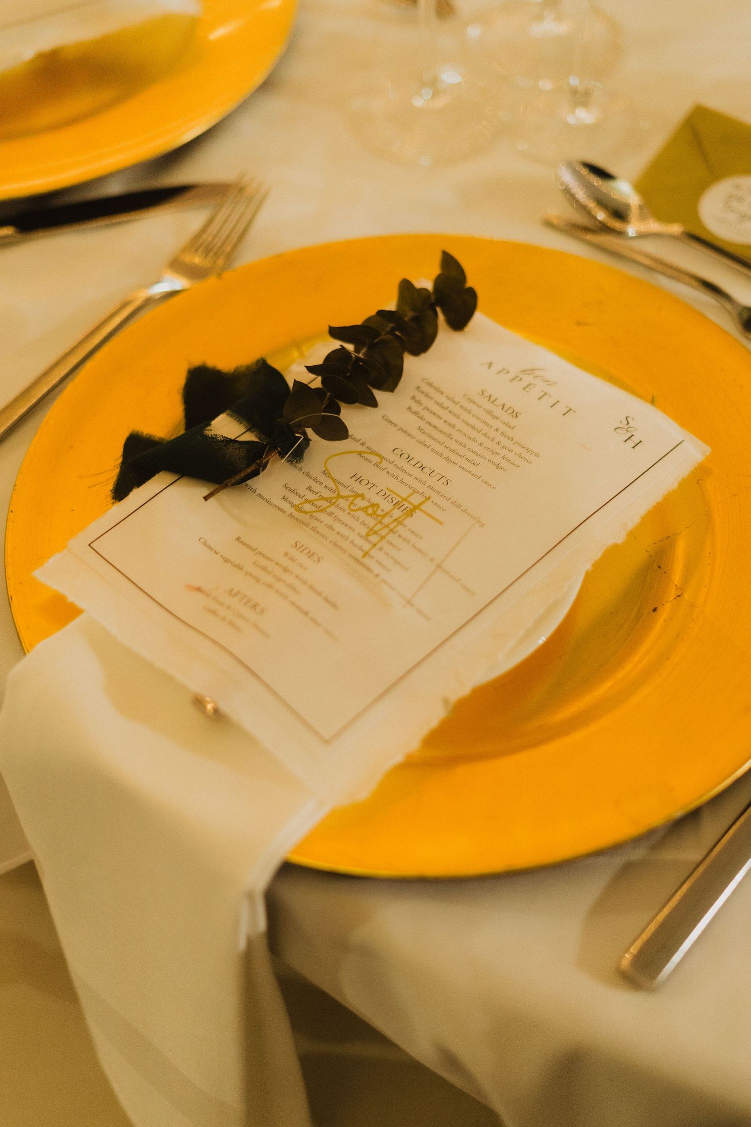 Gold charger plate with menu card and black leaf decoration on white tablecloth at formal place setting.