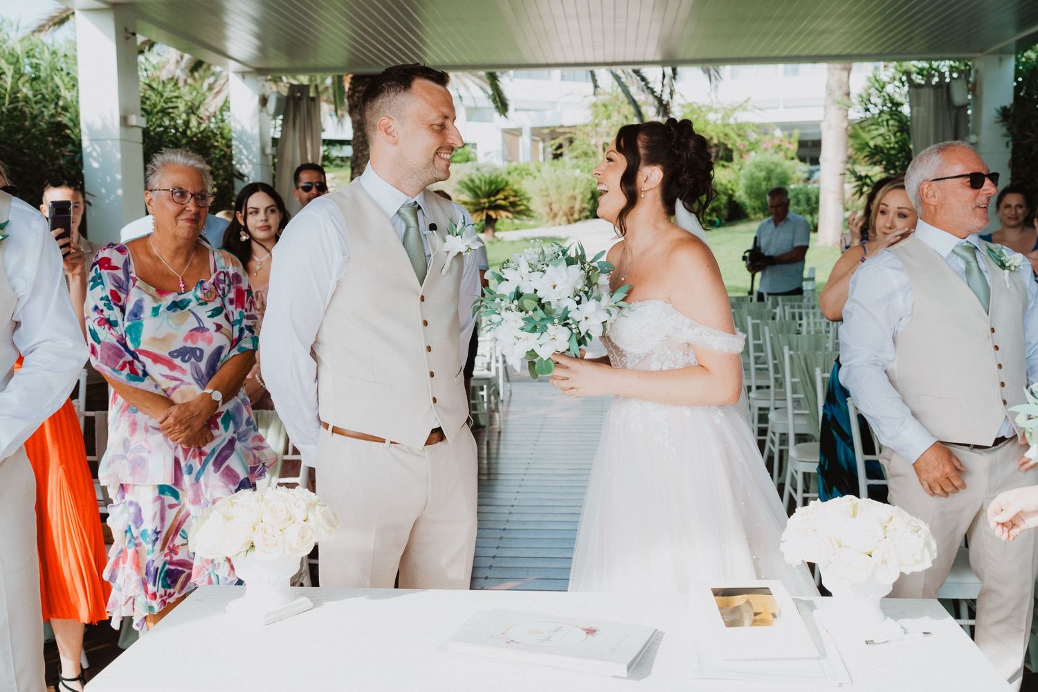A joyful outdoor wedding reception under a covered patio with guests in summer attire and floral decorations.