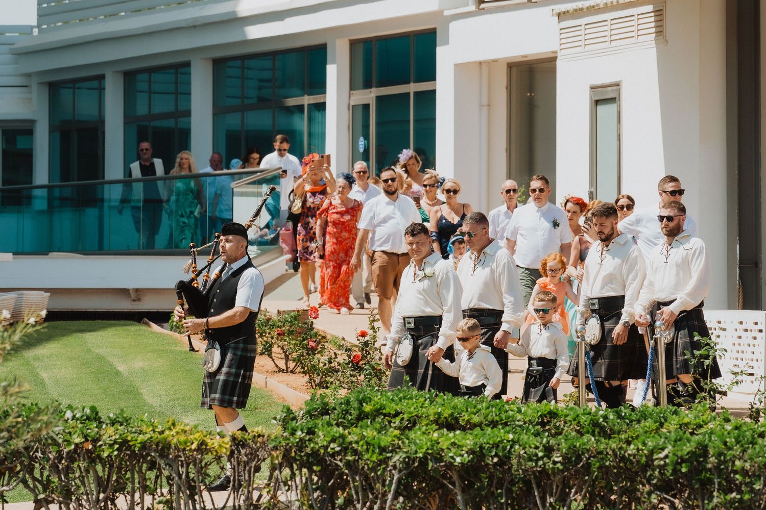 Wedding guests gather outdoors while bagpiper performs during ceremony.
