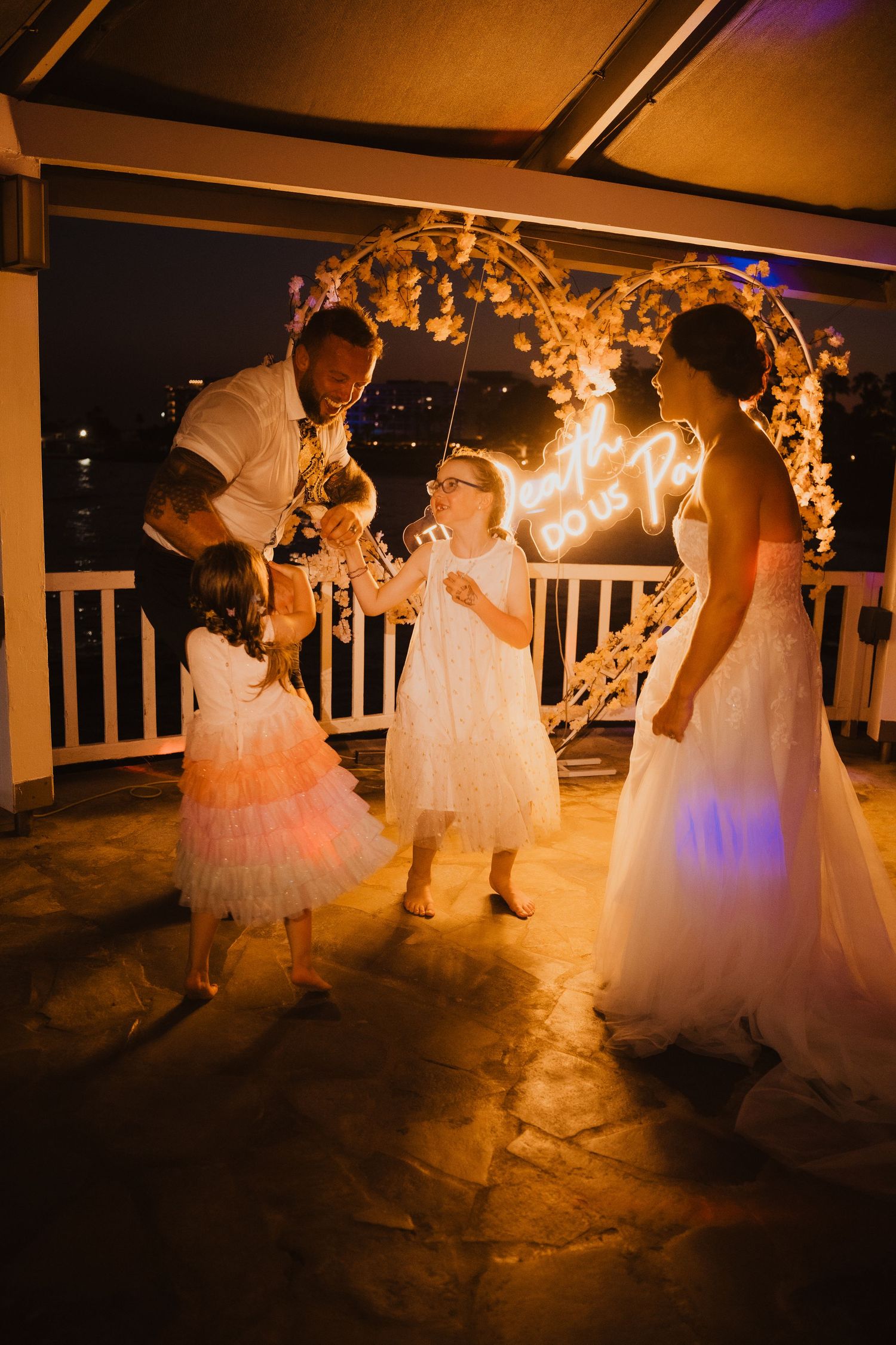 Wedding celebration under an illuminated floral arch on a seaside balcony at night.