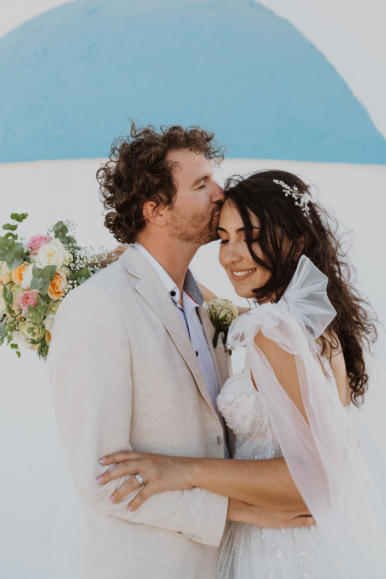 Tender wedding moment captured against white wall with blue dome in Greece.