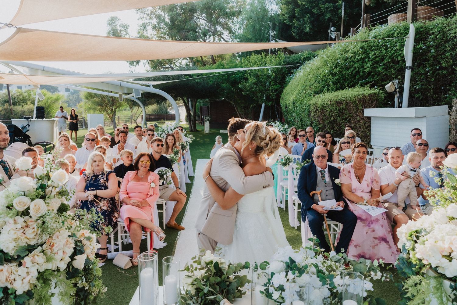 Outdoor garden wedding ceremony with white floral arrangements and guests seated under beige shade sails.