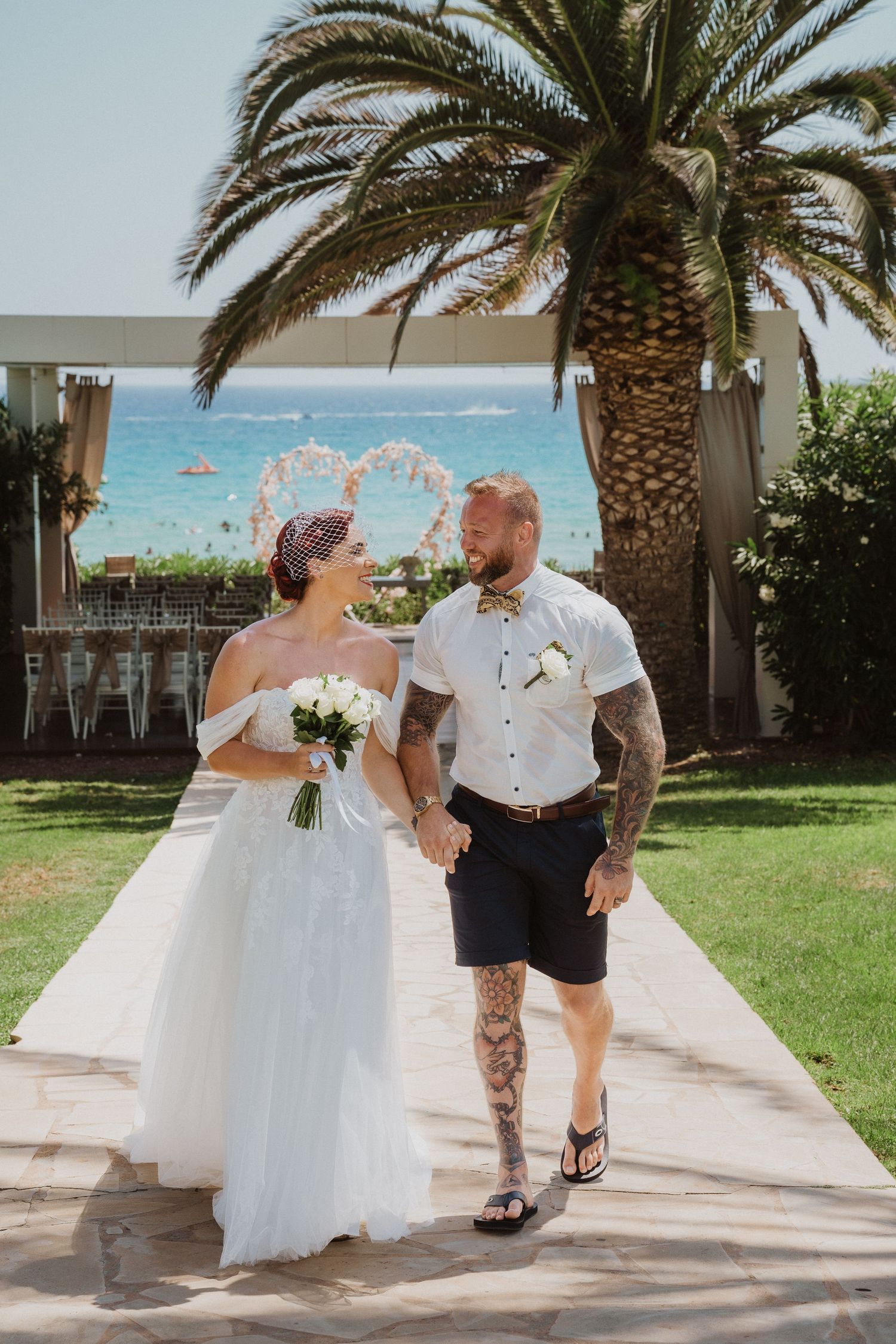 Couple walking together on stone path with palm trees and heart-shaped arch overlooking ocean view.