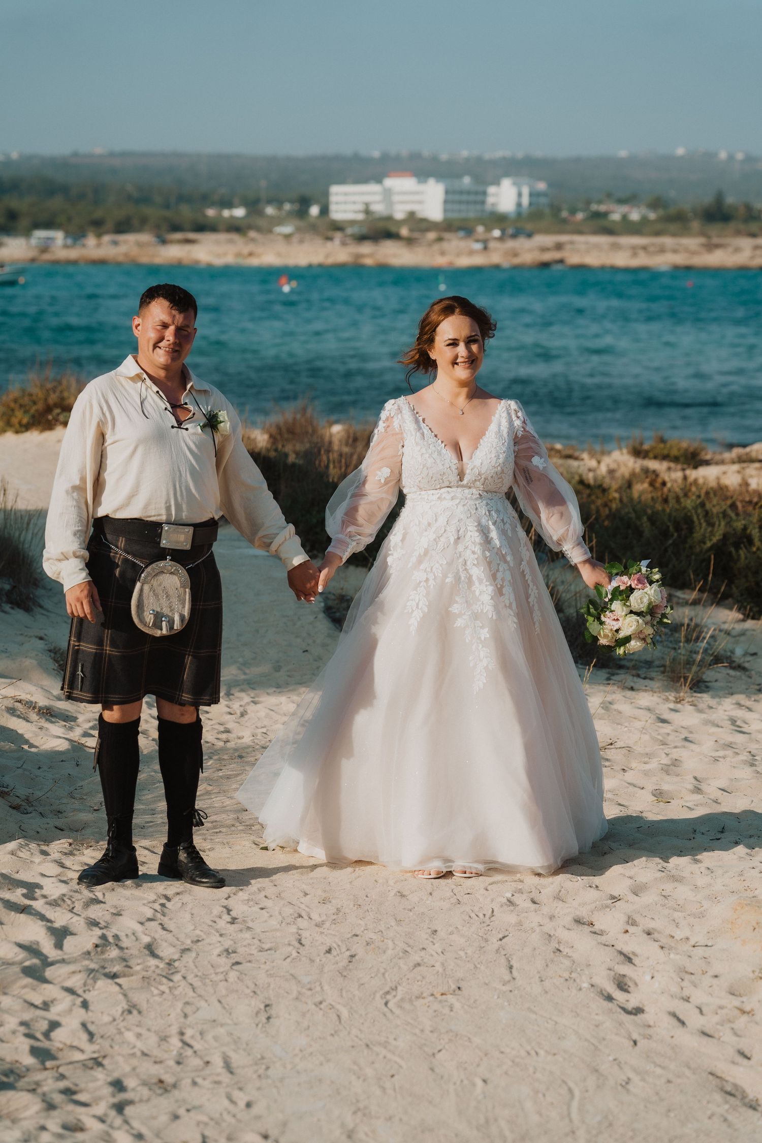 A beach wedding portrait featuring traditional Scottish attire and a lace wedding gown.