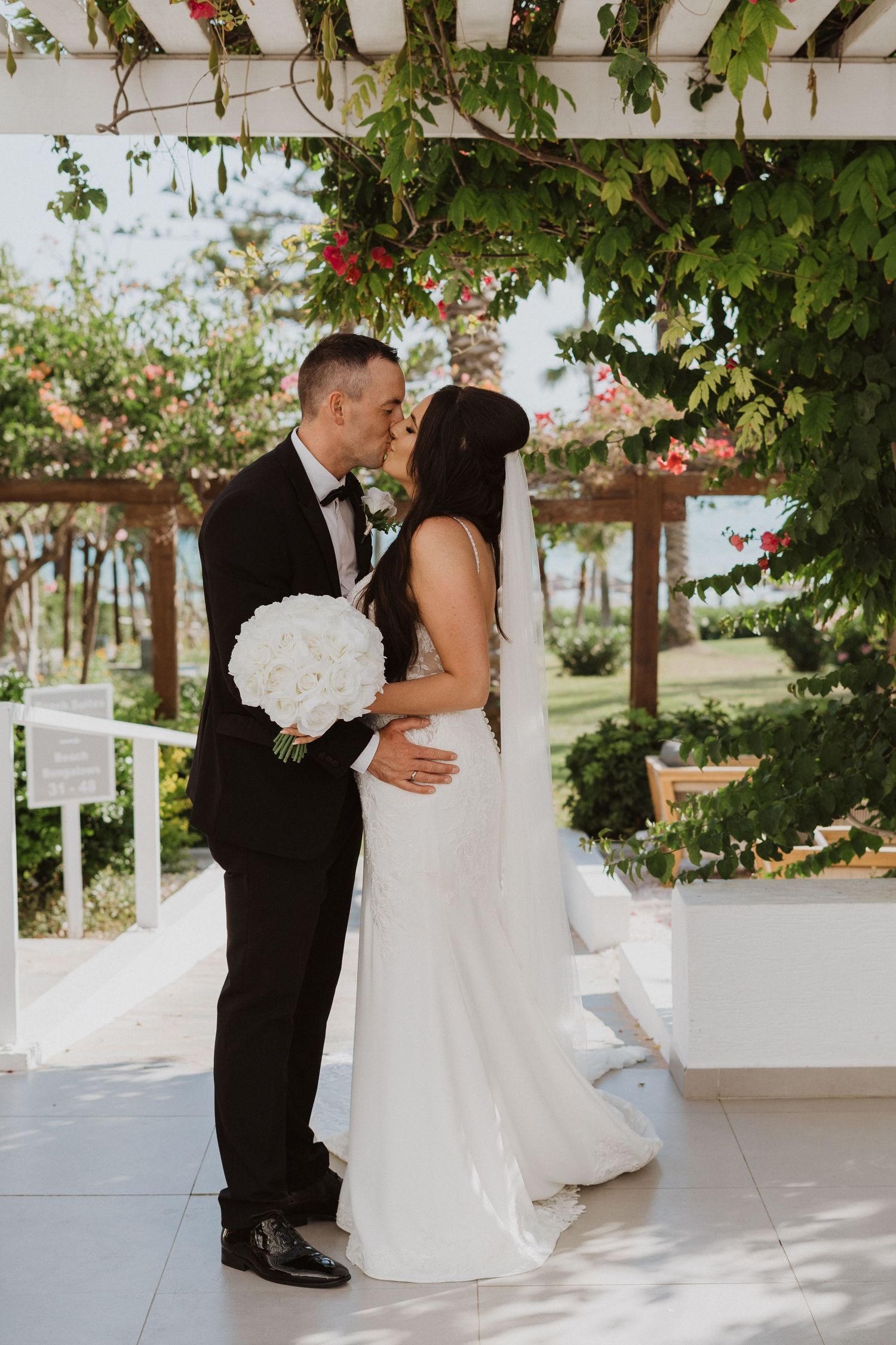 A couple embraces during their wedding celebration under a white pergola with tropical greenery.