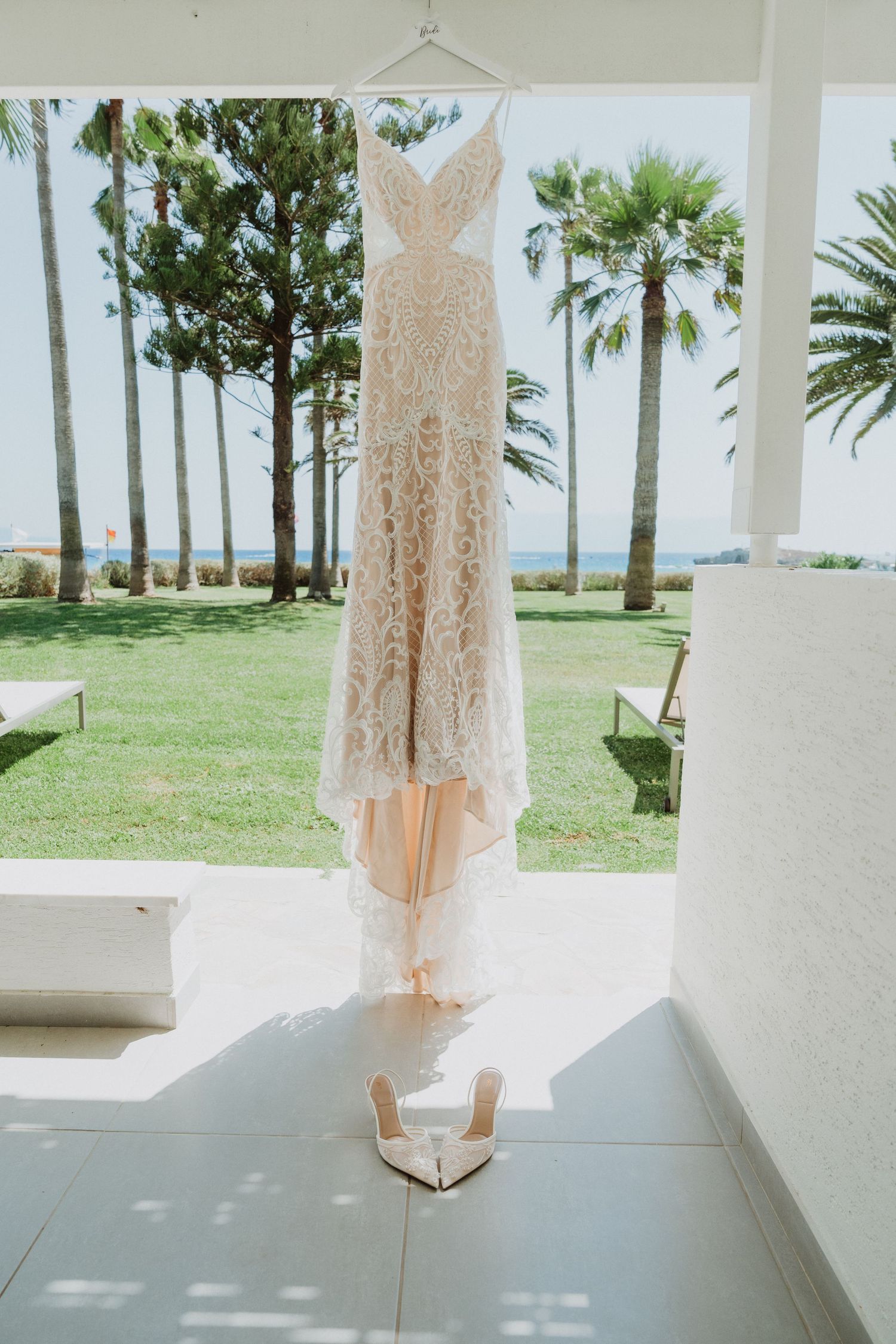 A delicate lace wedding dress hangs elegantly against a bright white wall with palm trees and ocean views visible outside.