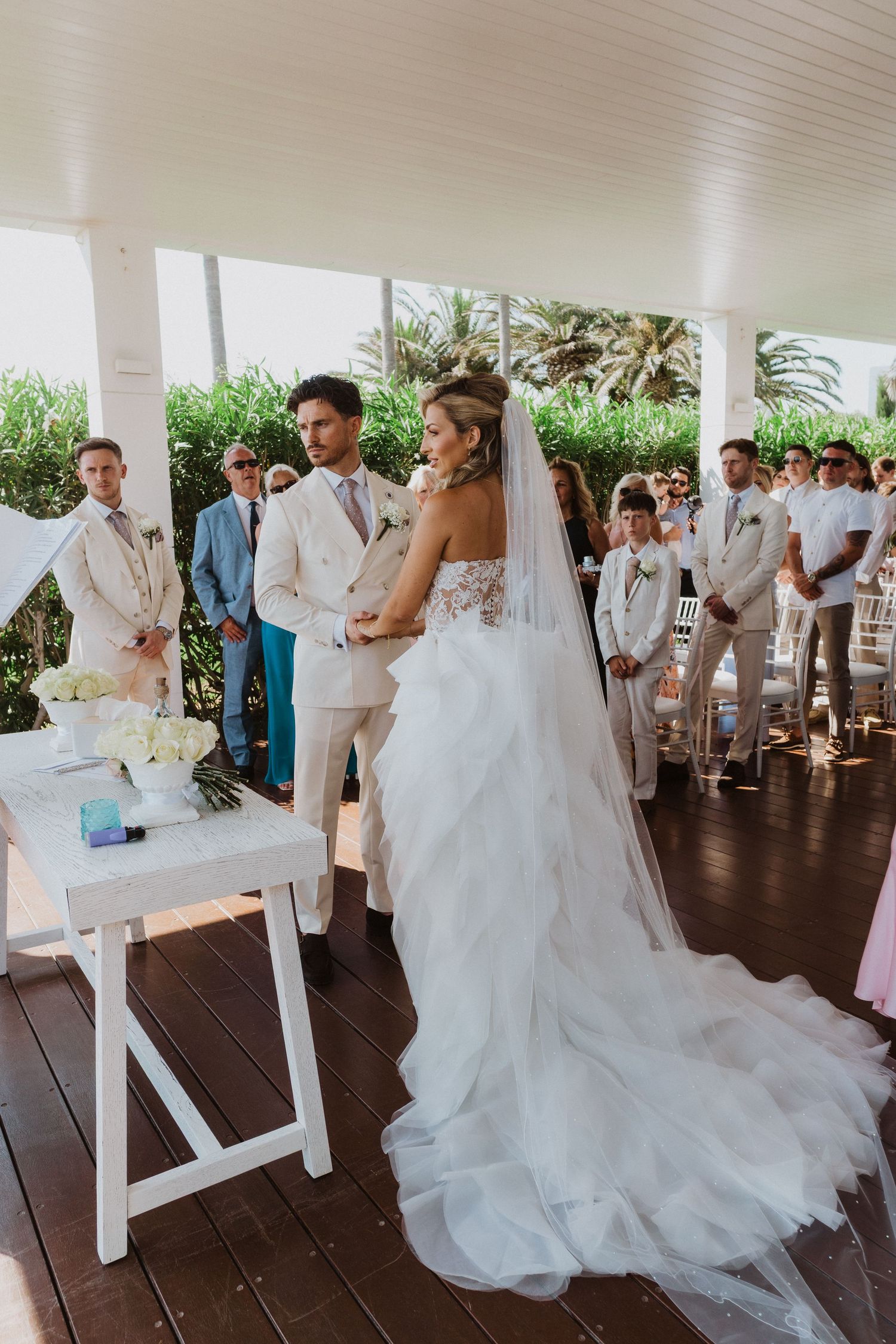 Wedding ceremony guests gathered on a covered outdoor patio with white chairs and elegant decor.