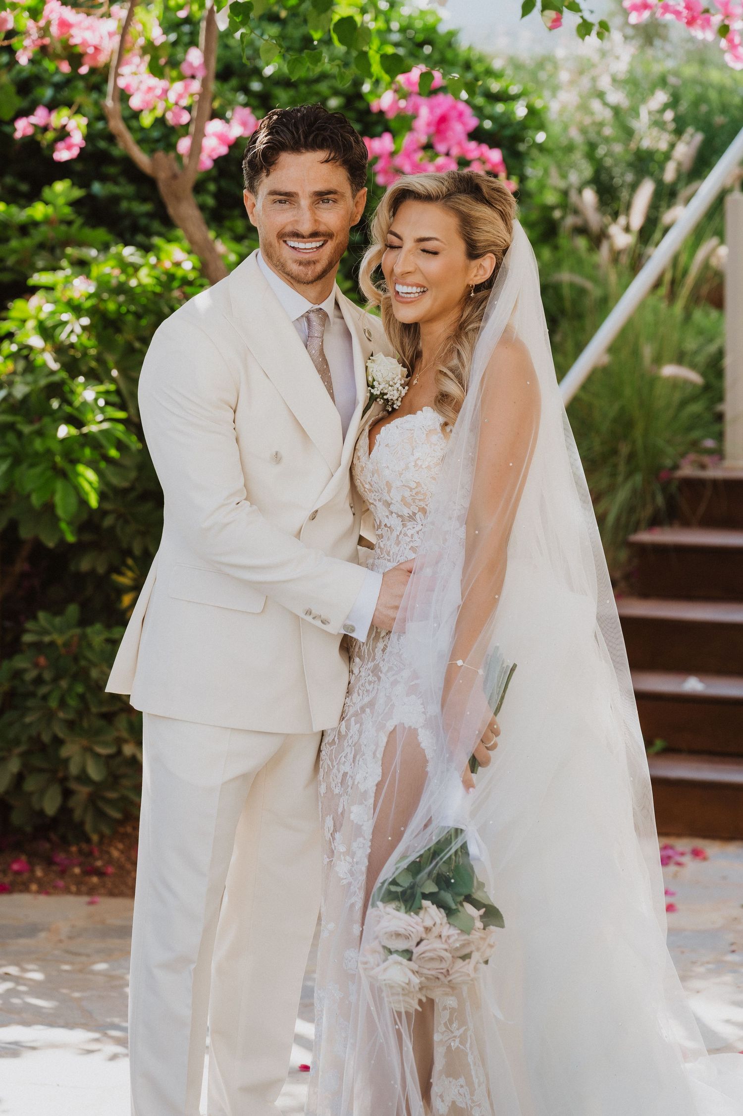 A wedding couple in white attire shares joyful moments together in a garden setting with pink flowers.