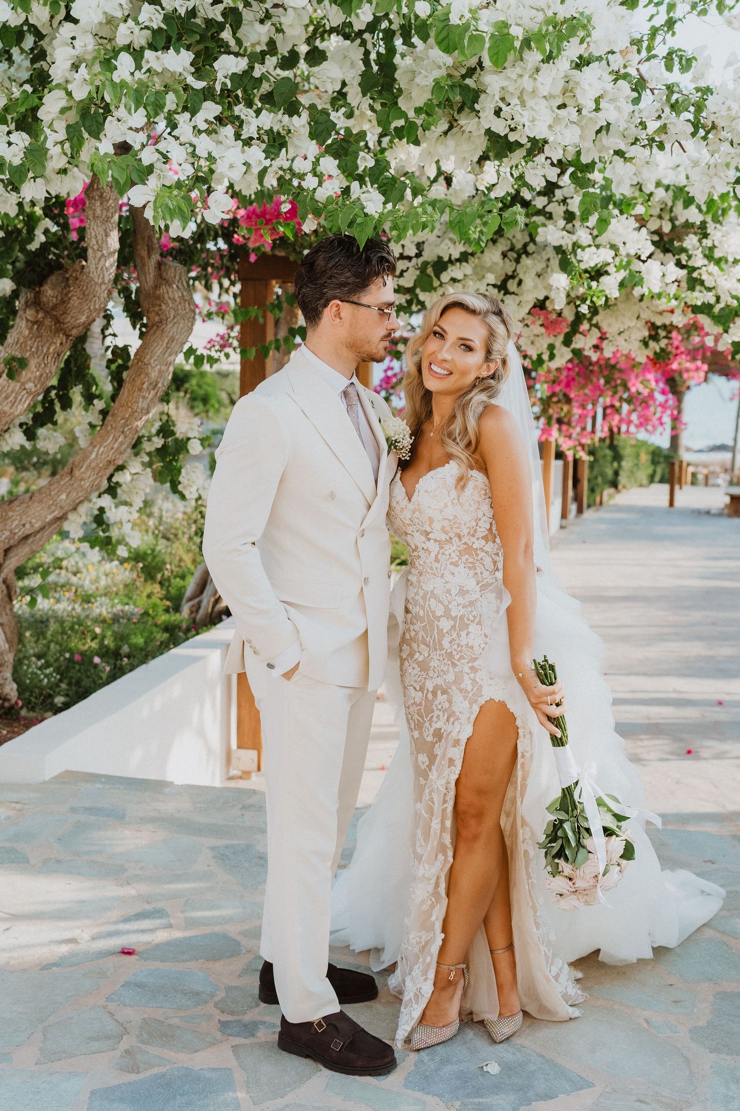 A couple poses under white flowering trees along a stone path in elegant wedding attire.