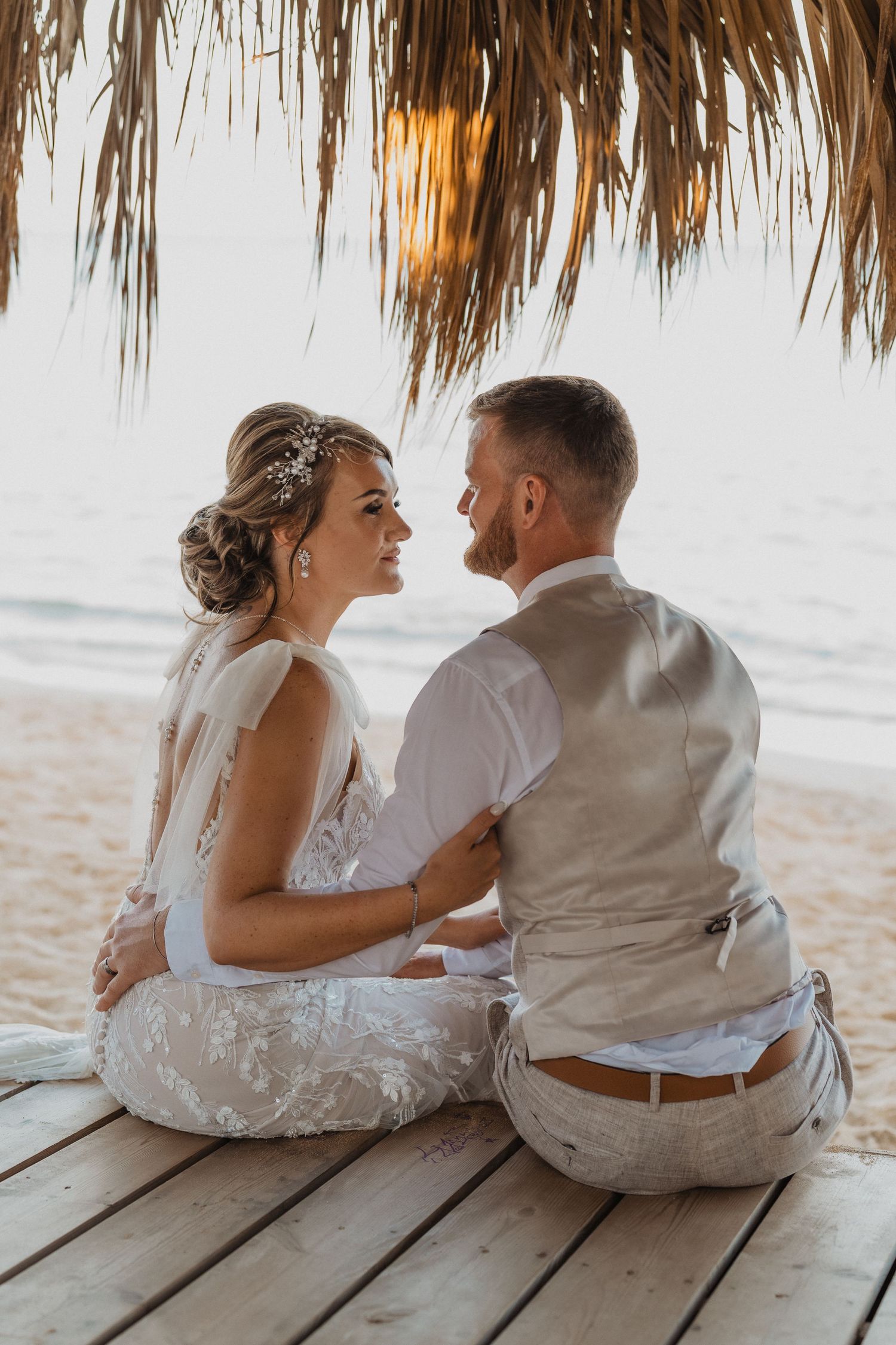 Couple sits together on wooden deck overlooking ocean under thatched roof.