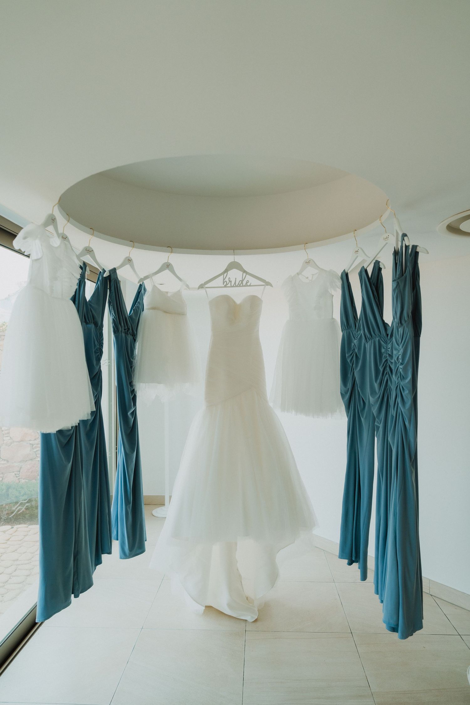 White wedding gown displayed on a bride hanger surrounded by teal bridesmaid dresses hanging in a bright circular alcove.