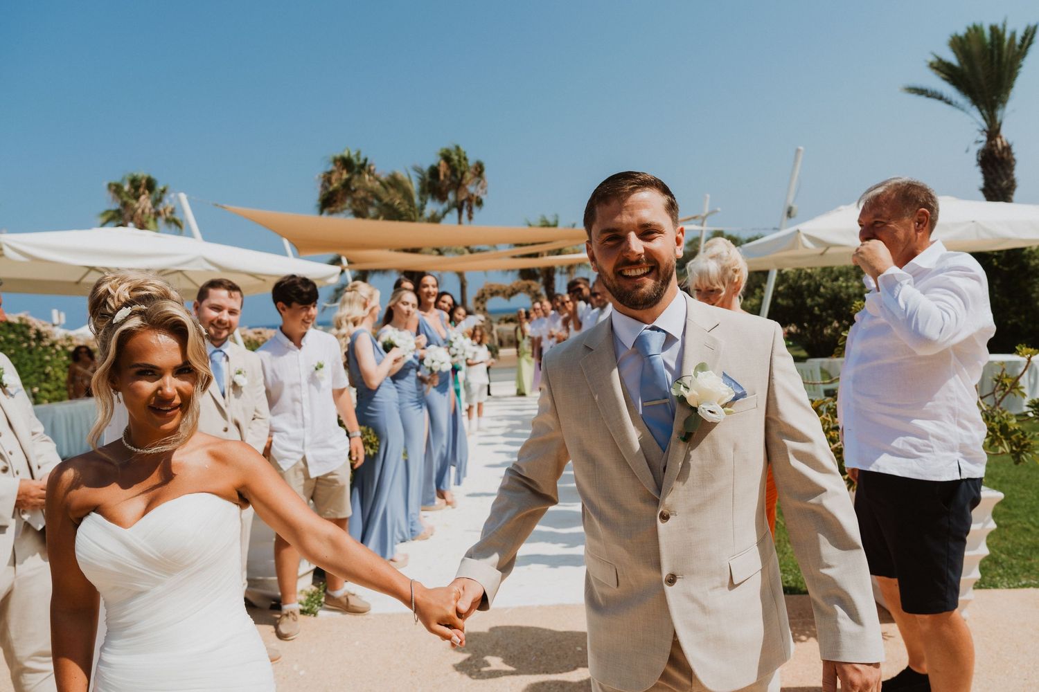 Bride and groom walk hand in hand after outdoor beach wedding ceremony with guests cheering in background.