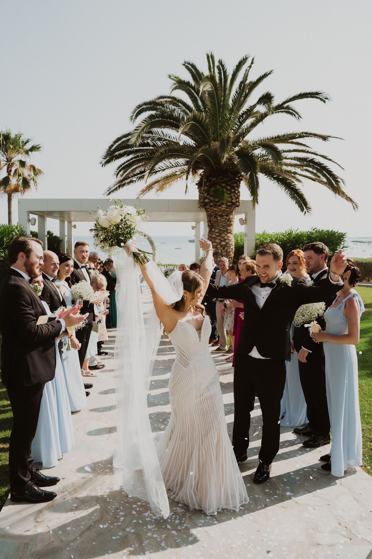 Joyful bride raises bouquet celebrating with groom and wedding party after outdoor ceremony near palm tree.