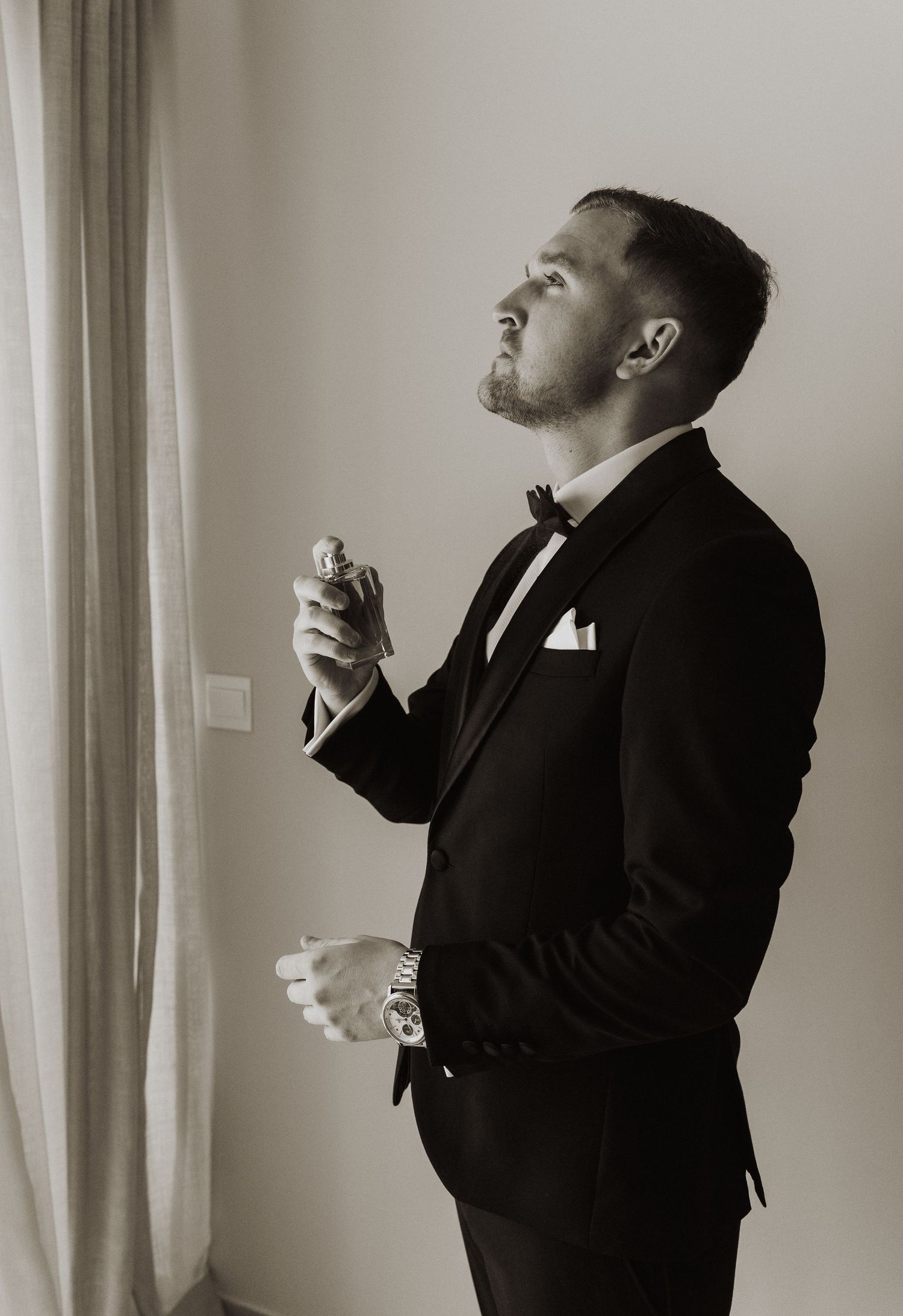 Dapper groom in black tuxedo applying cologne while getting ready for his wedding day.