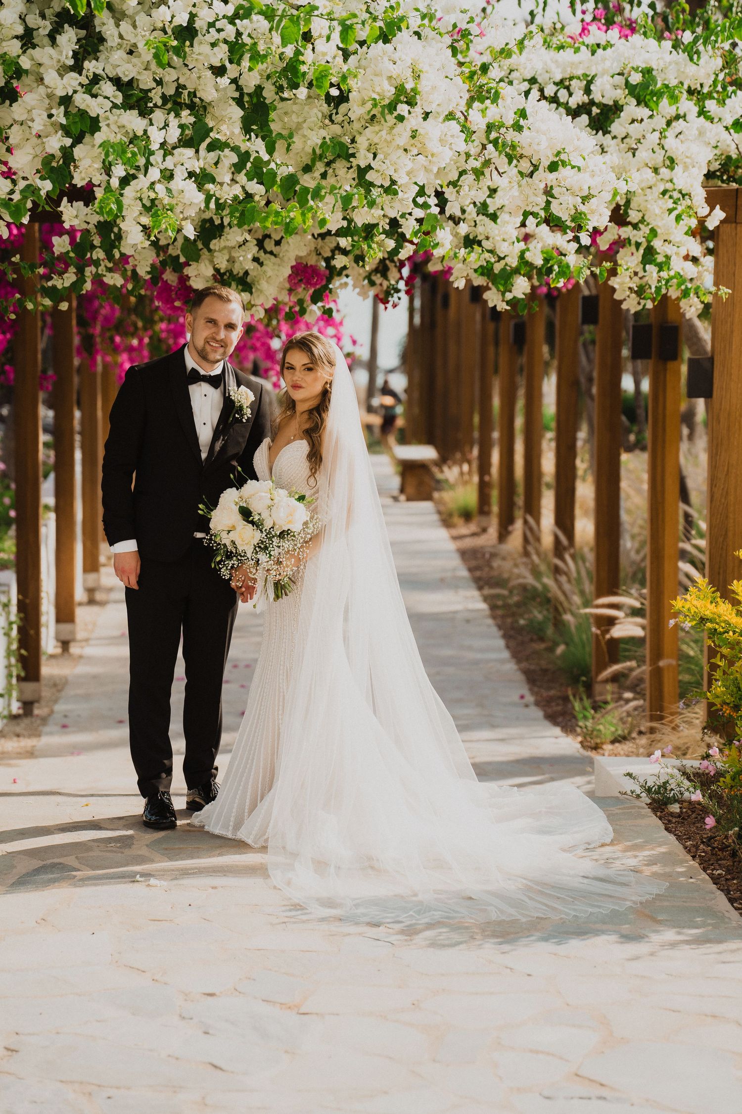 Bride and groom walk hand-in-hand under a stunning floral archway of white and pink blooms.