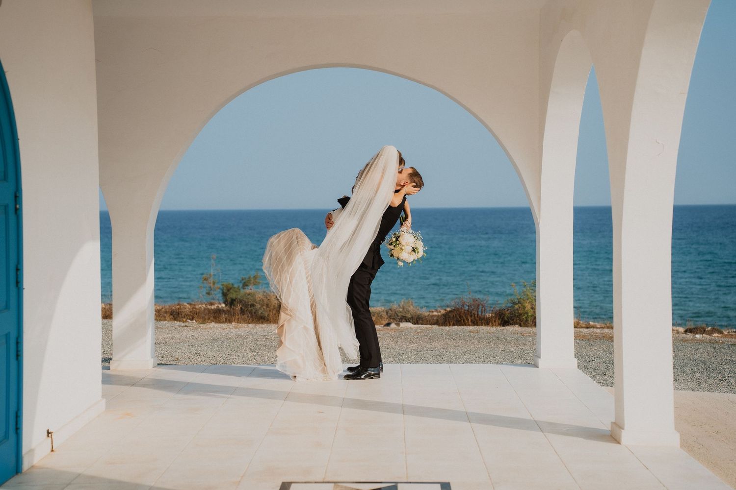 Groom dipping bride under a white archway with stunning blue ocean views in the background.