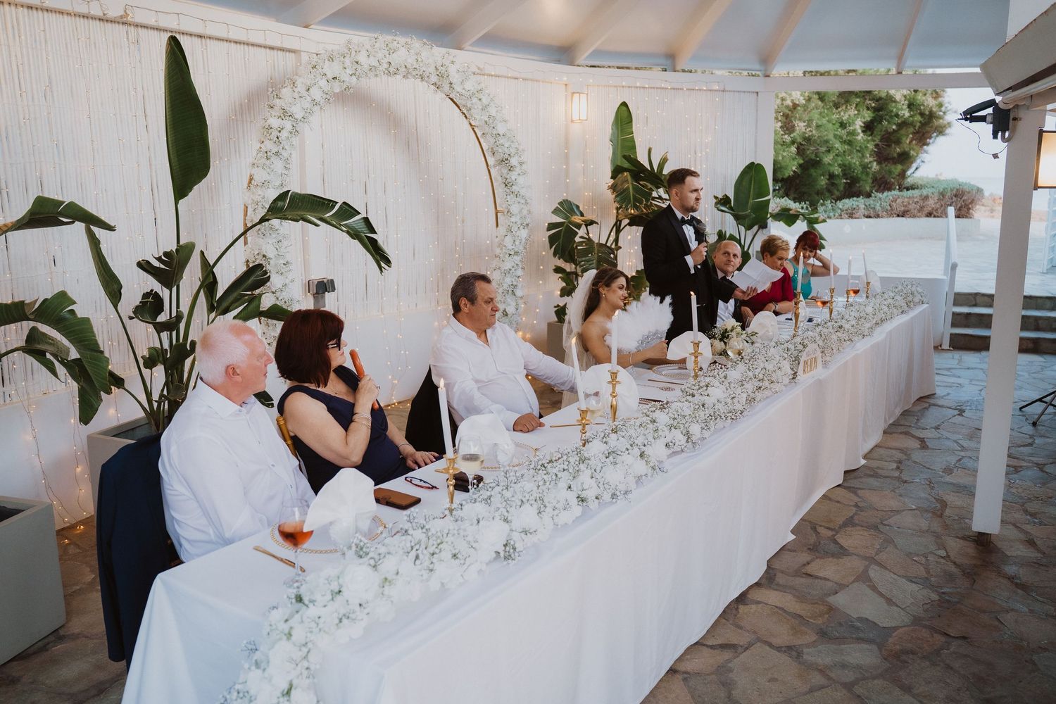Wedding party seated at a long white floral head table during outdoor reception under a white canopy.