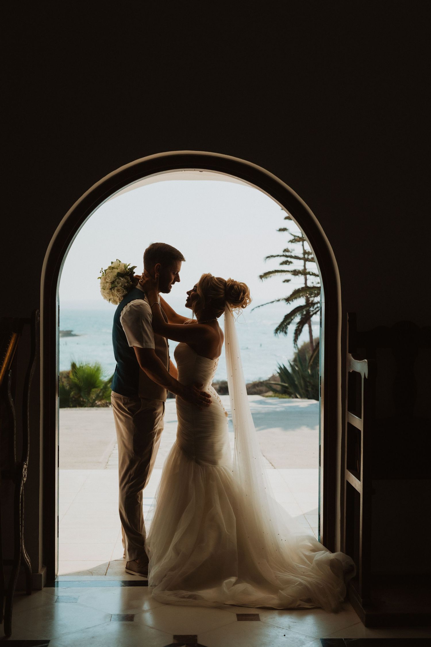Silhouette of bride and groom embracing in arched doorway with ocean view backdrop on their wedding day.