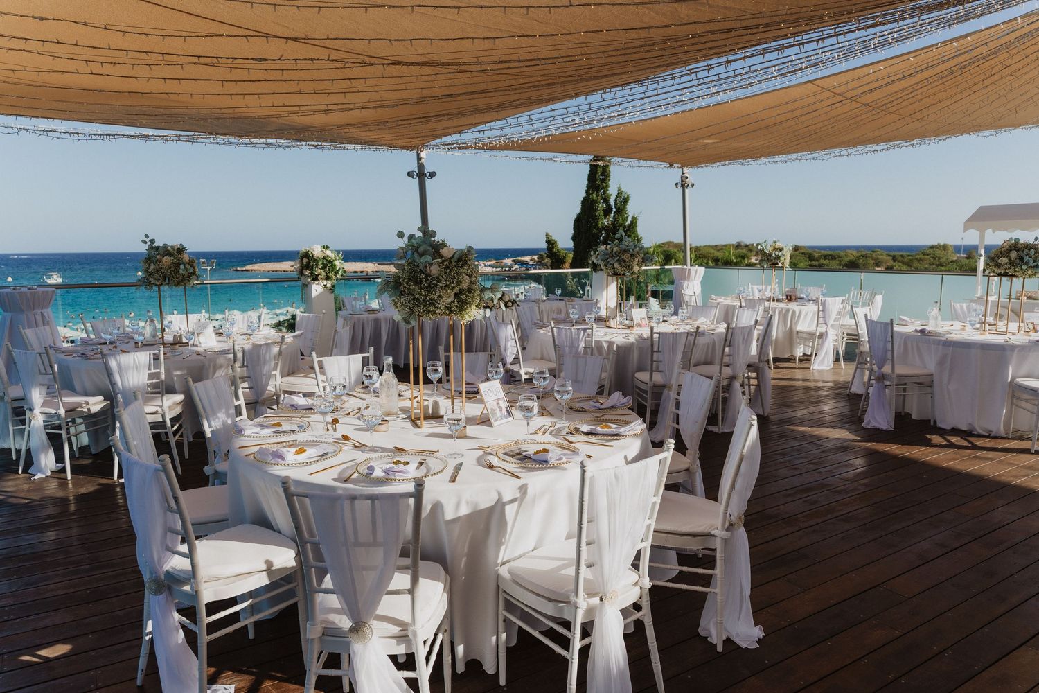 Elegant wedding reception setup with round tables and white linens overlooking an oceanfront venue at sunset.