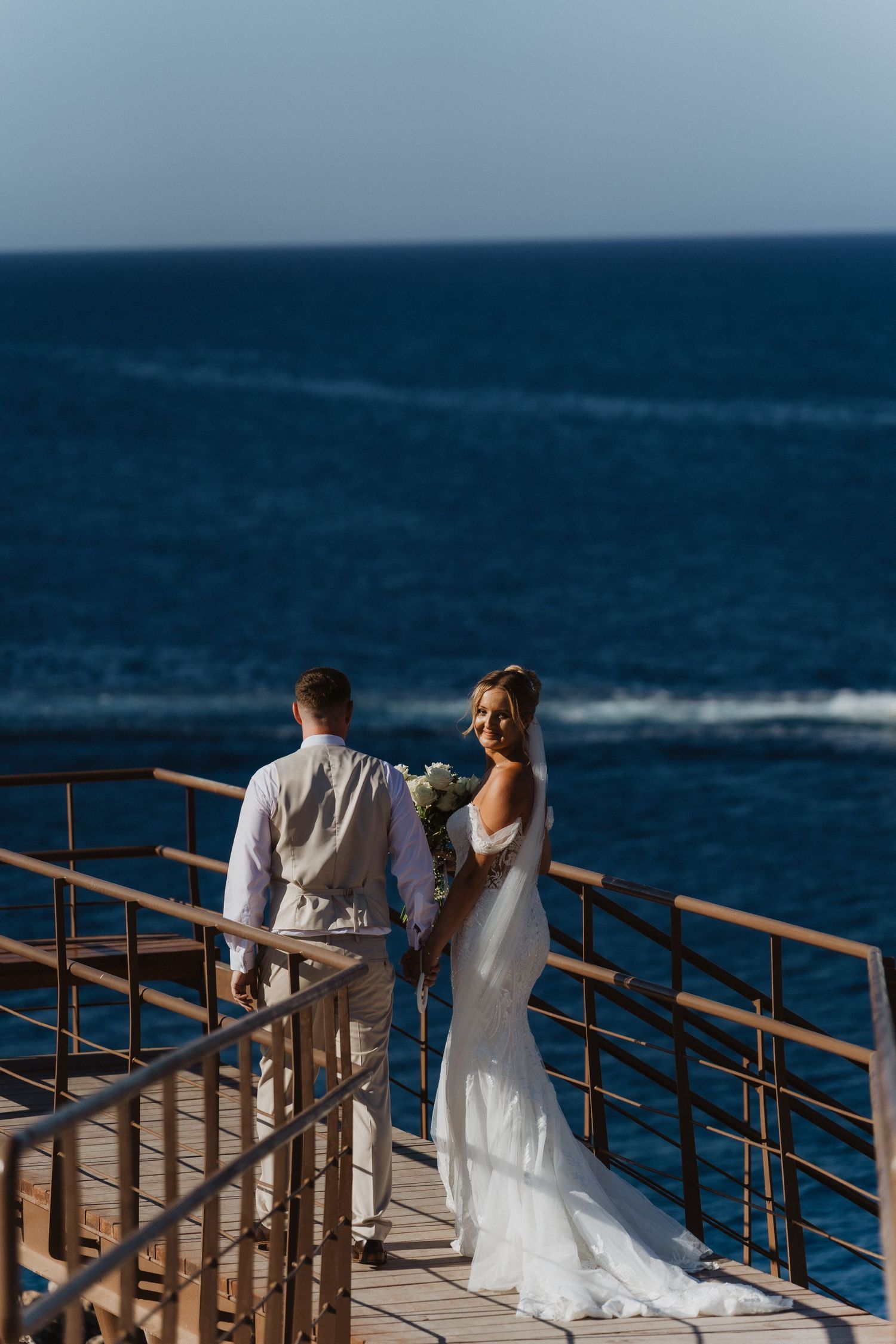 A couple stands on an oceanfront deck during their destination wedding, with the bride wearing a flowing white gown.
