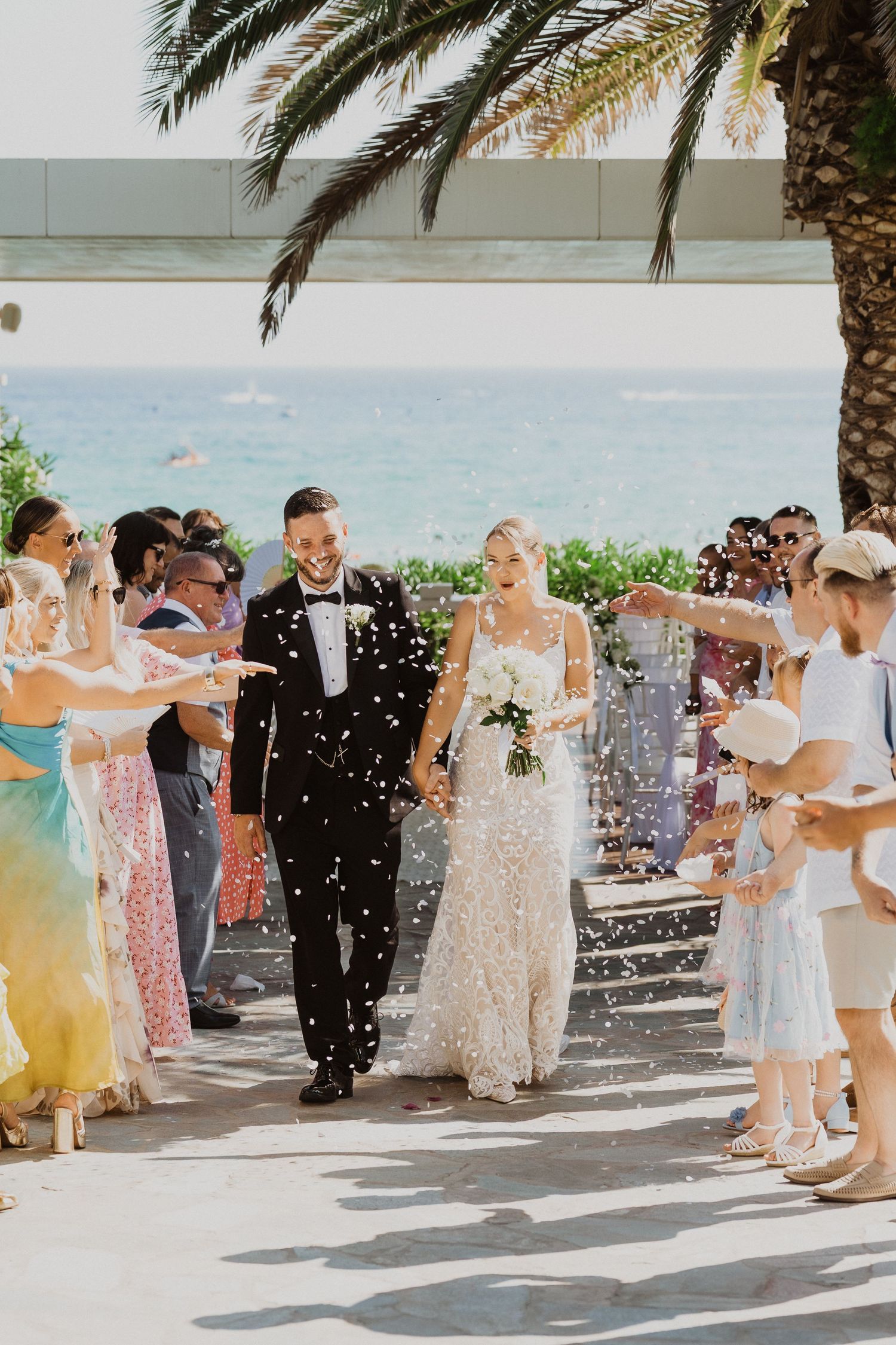 Wedding guests celebrate with confetti under palm trees at a beachside venue.