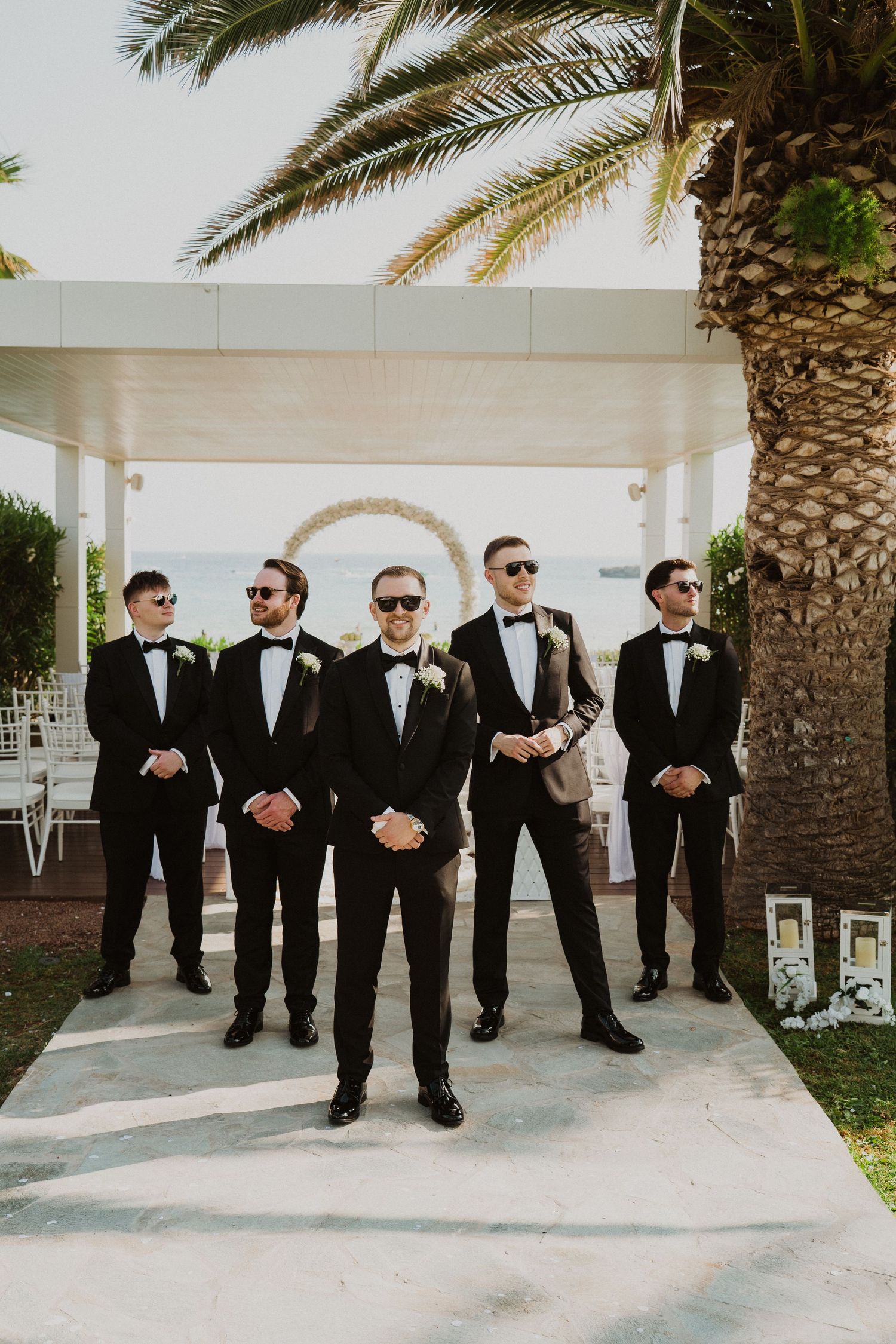 Groom and groomsmen in black tuxedos with sunglasses pose confidently at outdoor wedding ceremony.