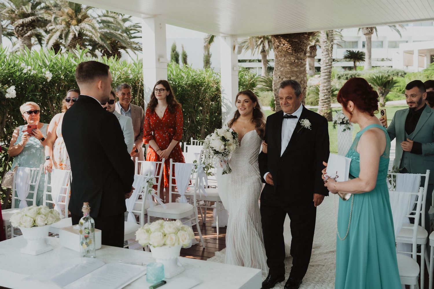 Bride walking down aisle with father toward groom during outdoor wedding ceremony under pavilion.