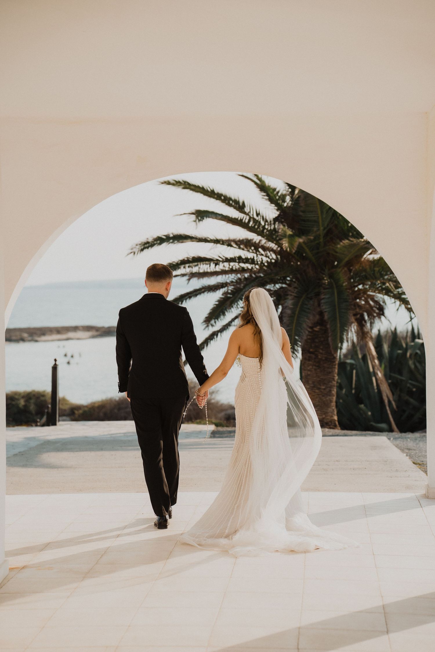 Bride and groom holding hands walking through a circular arch with palm tree and ocean view beyond.