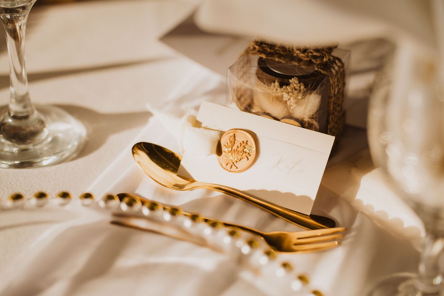 Elegant gold cutlery, wax seal place card, and small gift box on a white wedding table setting.