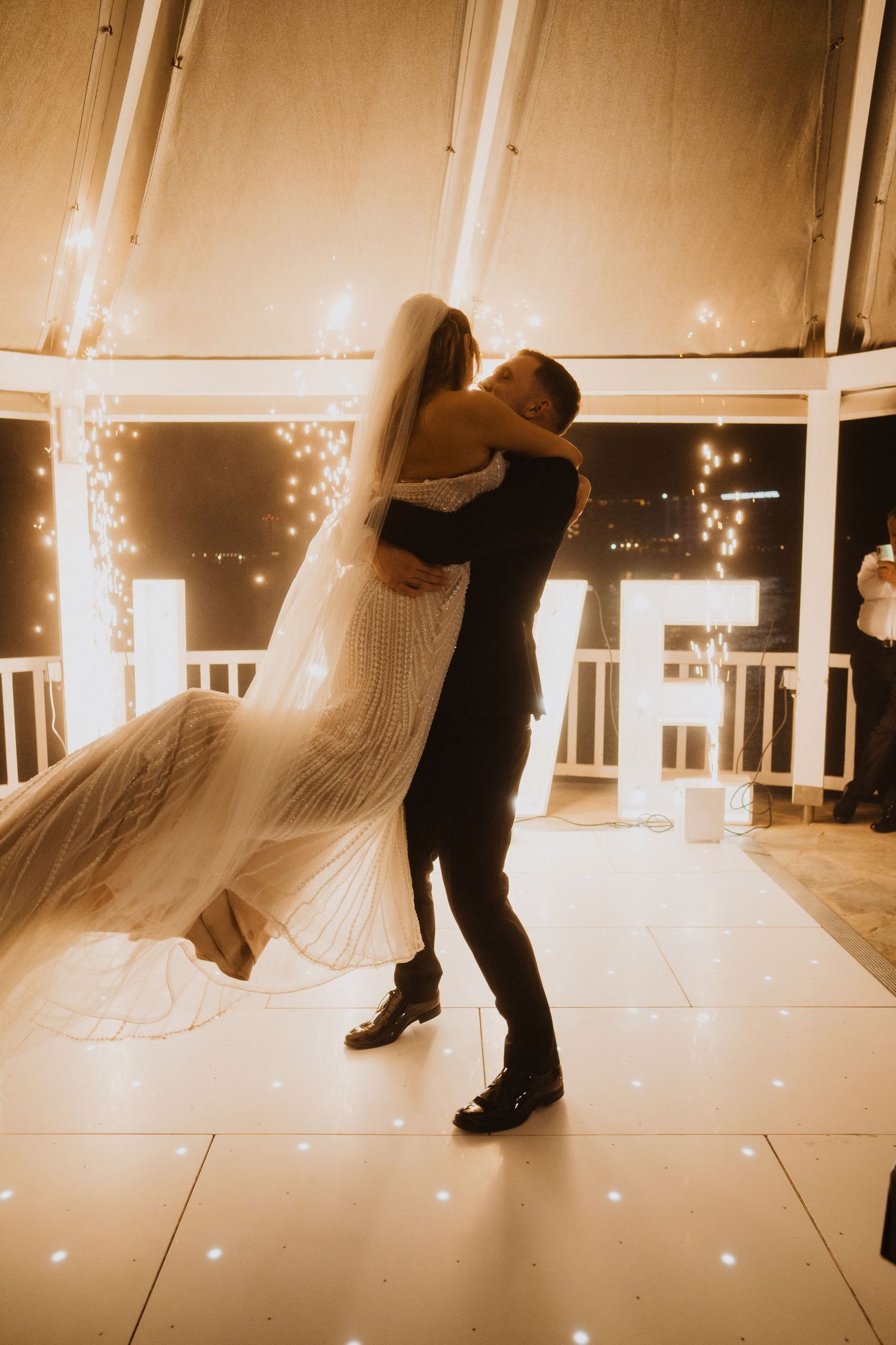 Groom lifts bride in wedding gown on a lit dance floor with LOVE letters glowing in the background.