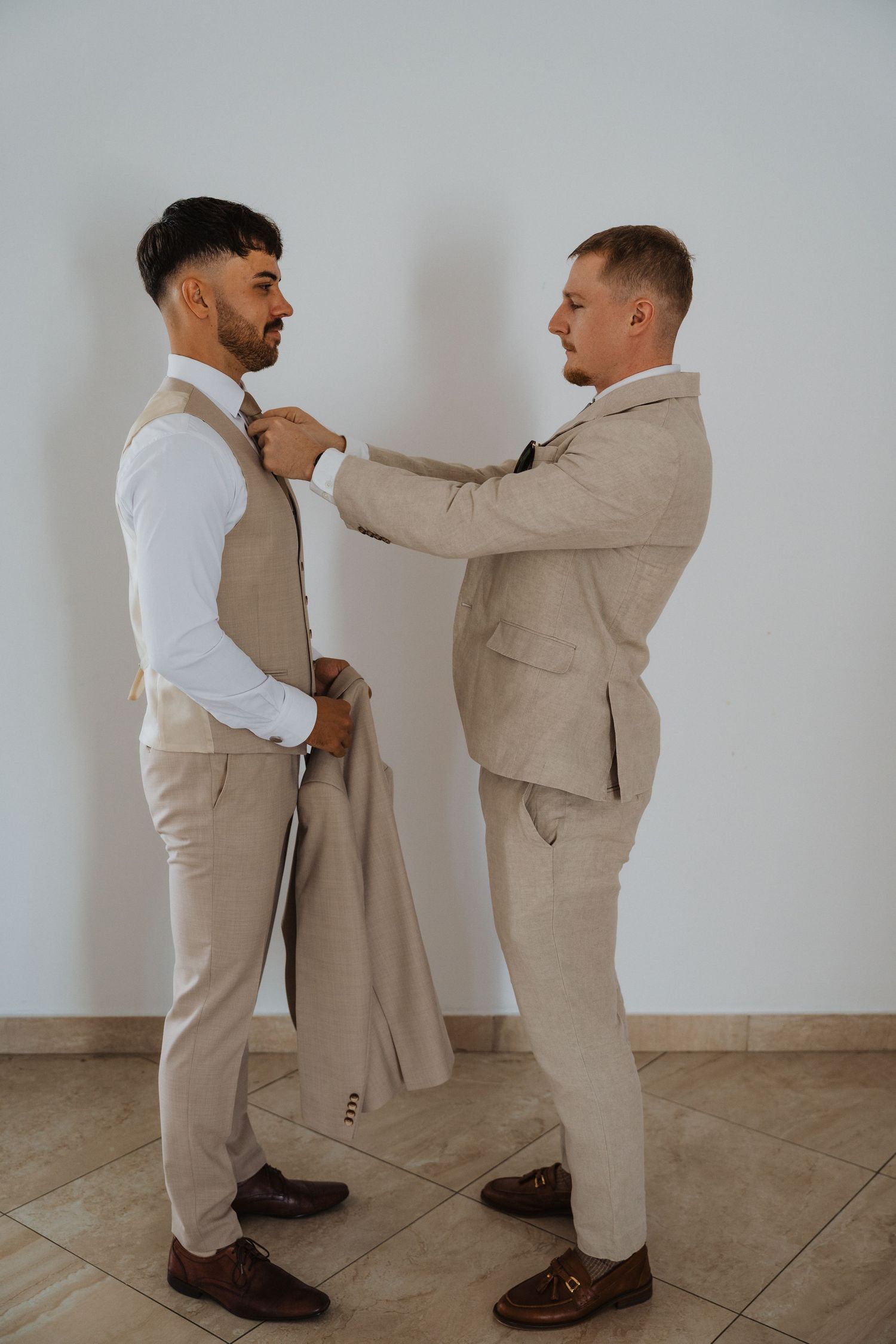 Two groomsmen in beige suits helping each other get ready for a wedding.