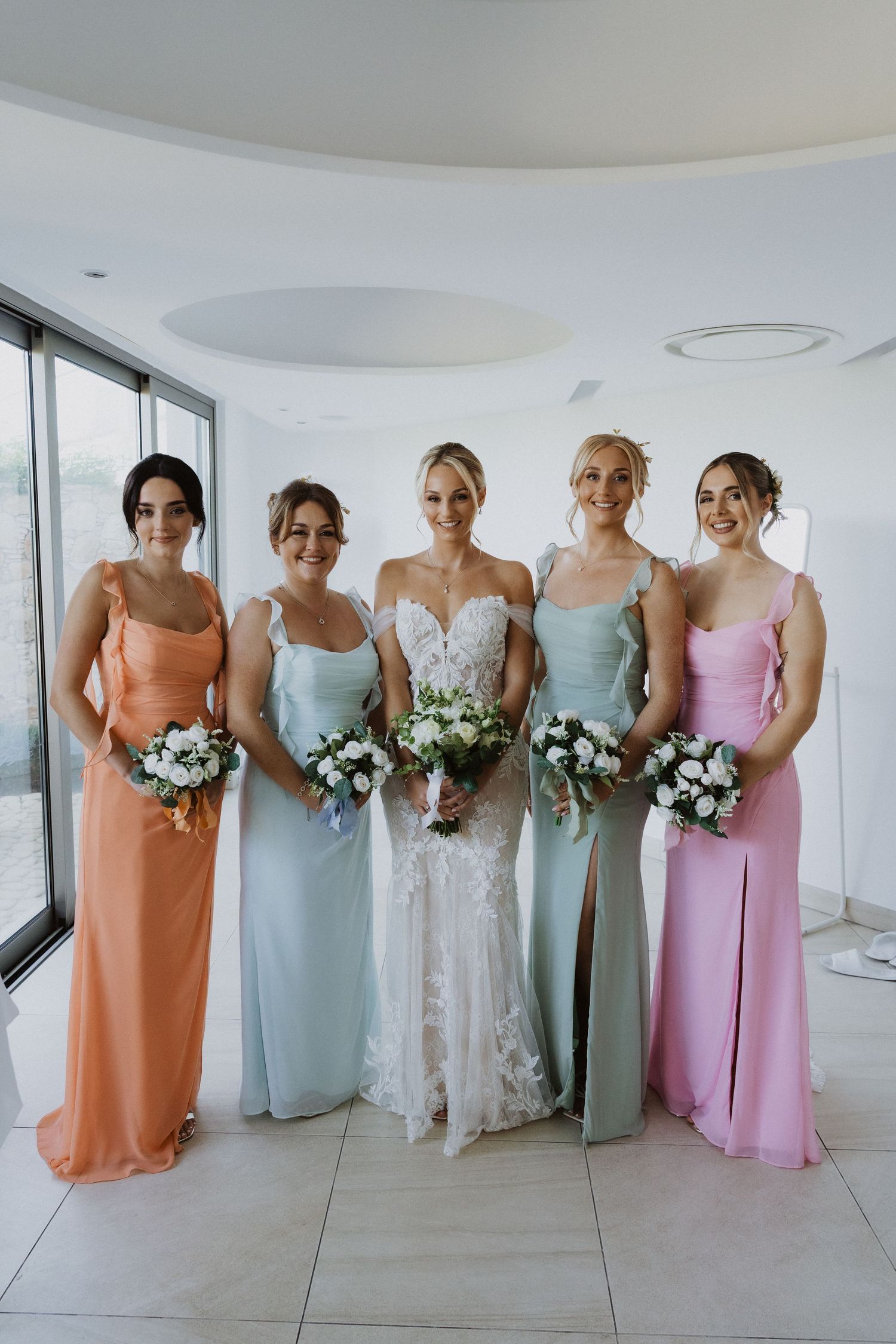Bridesmaids in pastel dresses holding white bouquets pose together in a modern white venue.