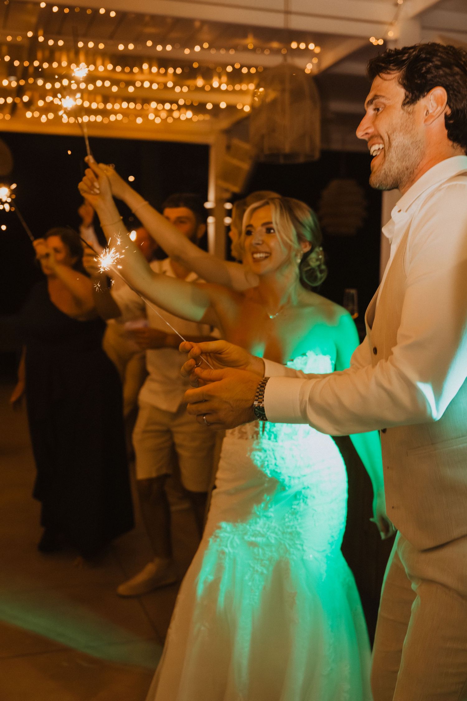 Wedding celebration with dancing and sparklers under string lights and green ambient glow.
