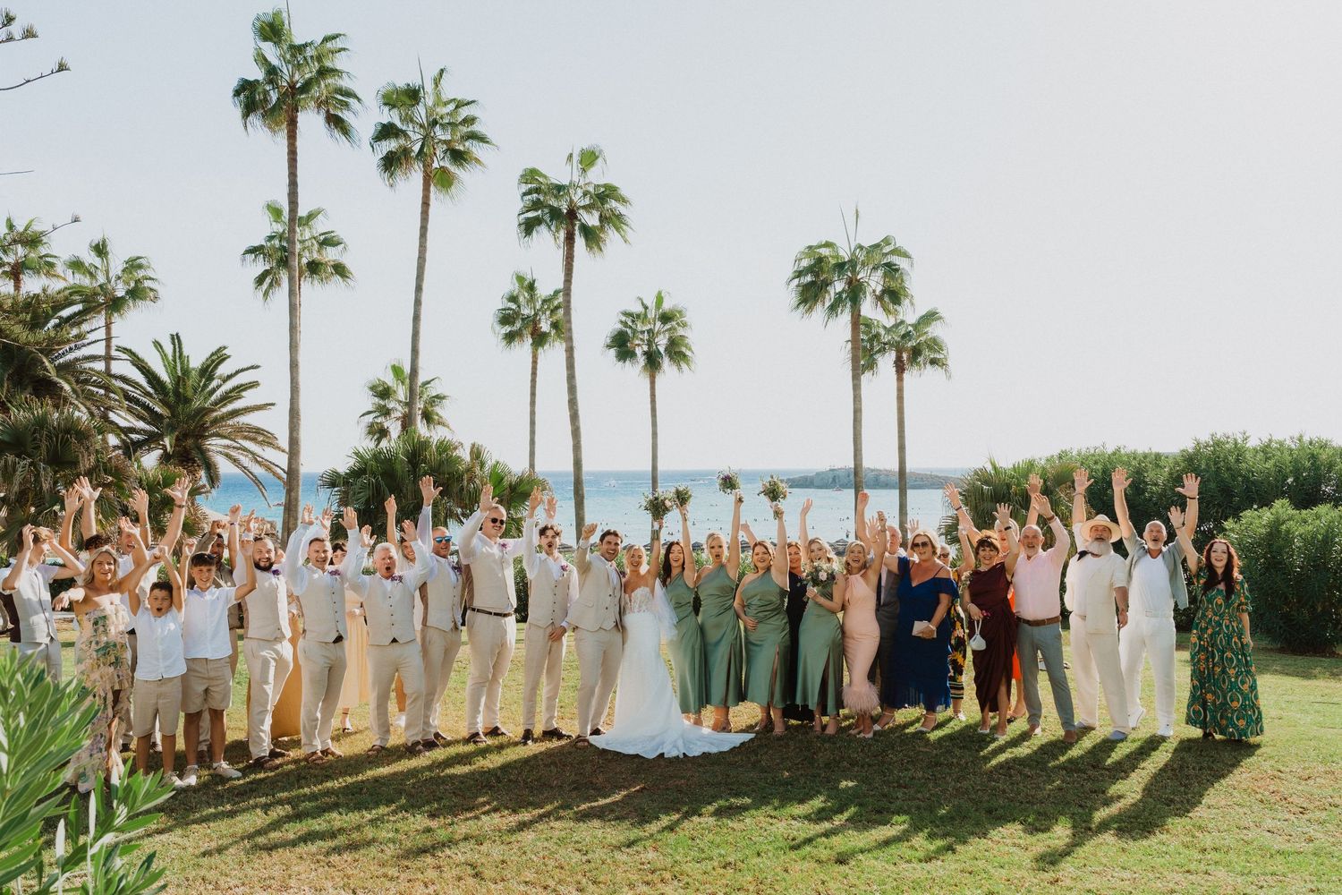 Large wedding party group photo with palm trees and ocean view in the background.