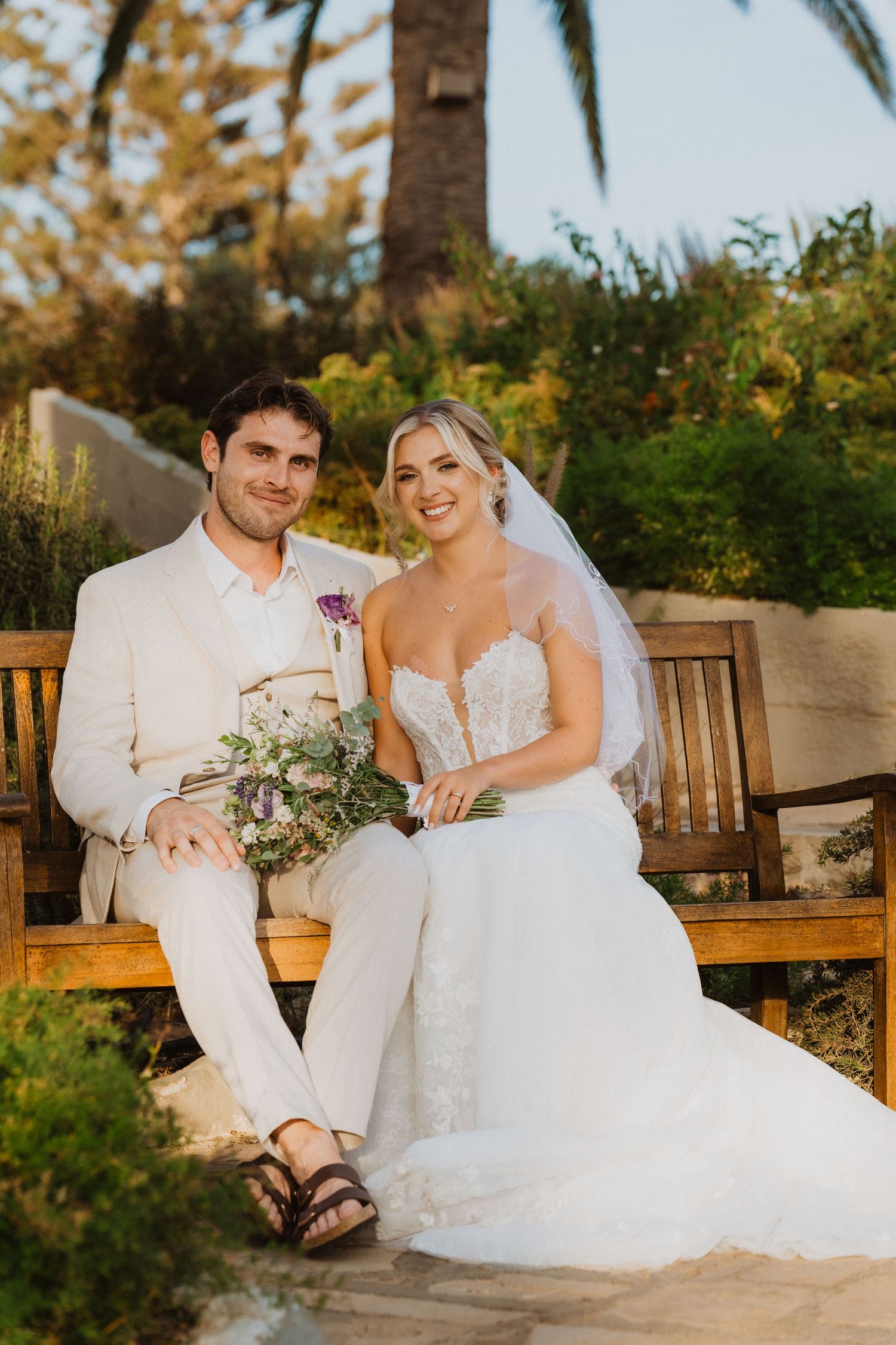 Newlyweds relax on a wooden bench in a tropical garden setting.