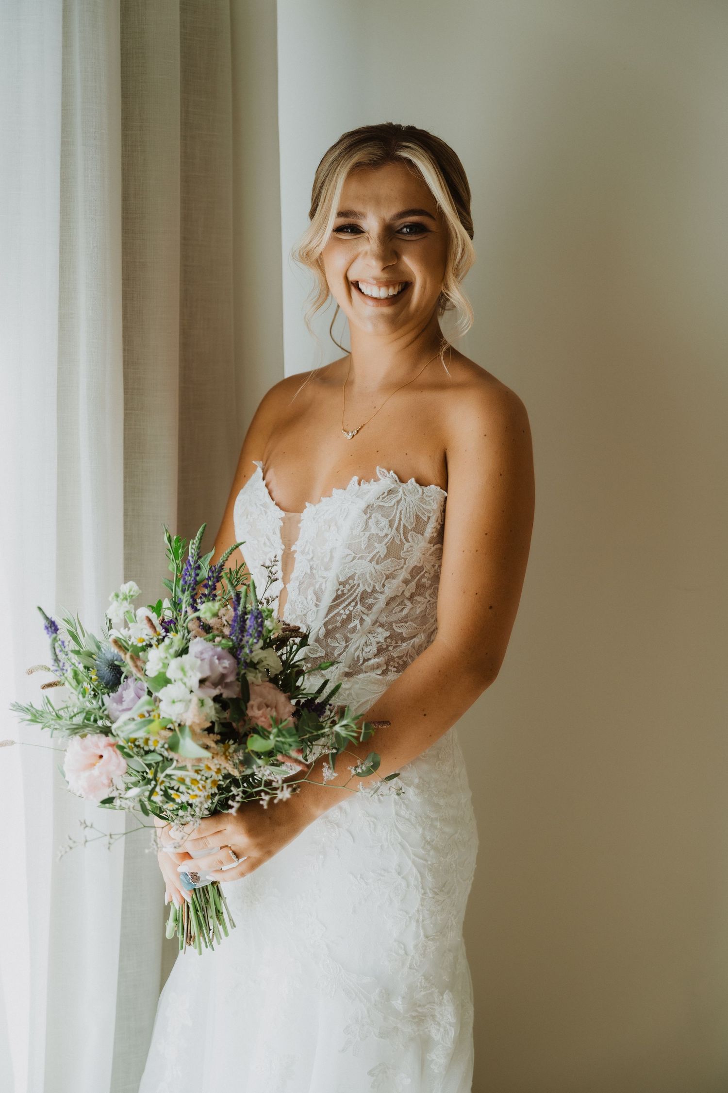 Joyful bride in strapless lace gown holding a purple and white wildflower bouquet.
