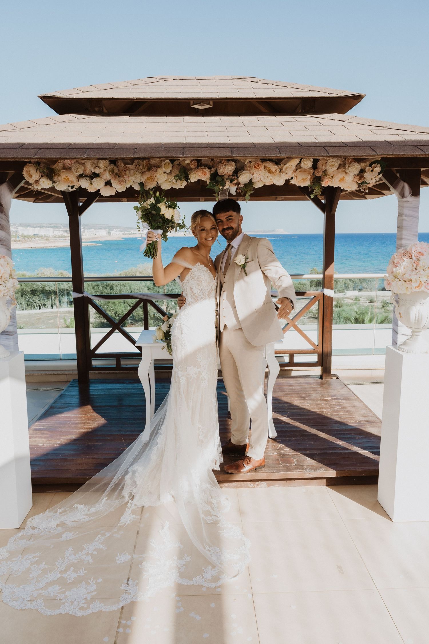 A wedding couple poses in a beachside gazebo decorated with white flowers and ocean views.