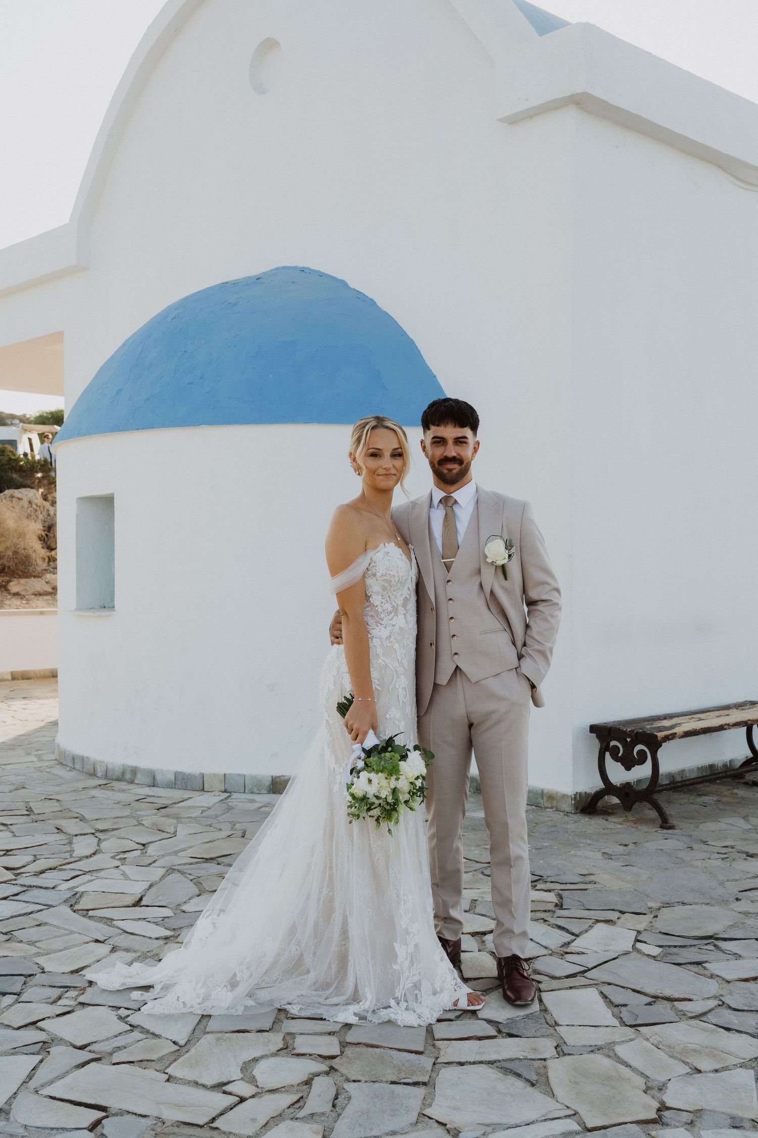 Wedding couple poses in front of traditional white Greek church with blue dome.