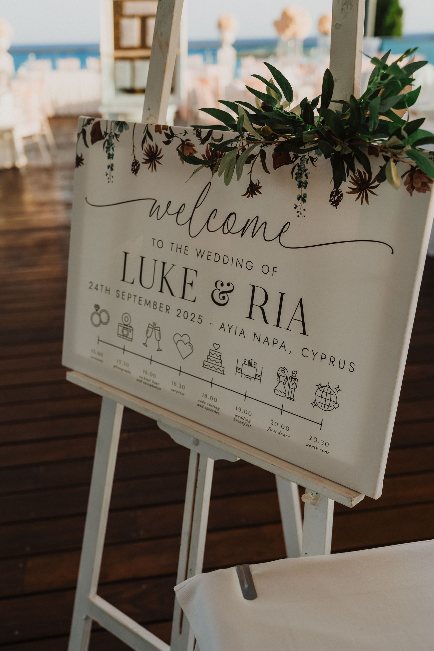 Wedding welcome sign with floral design and timeline of events displayed on white easel.